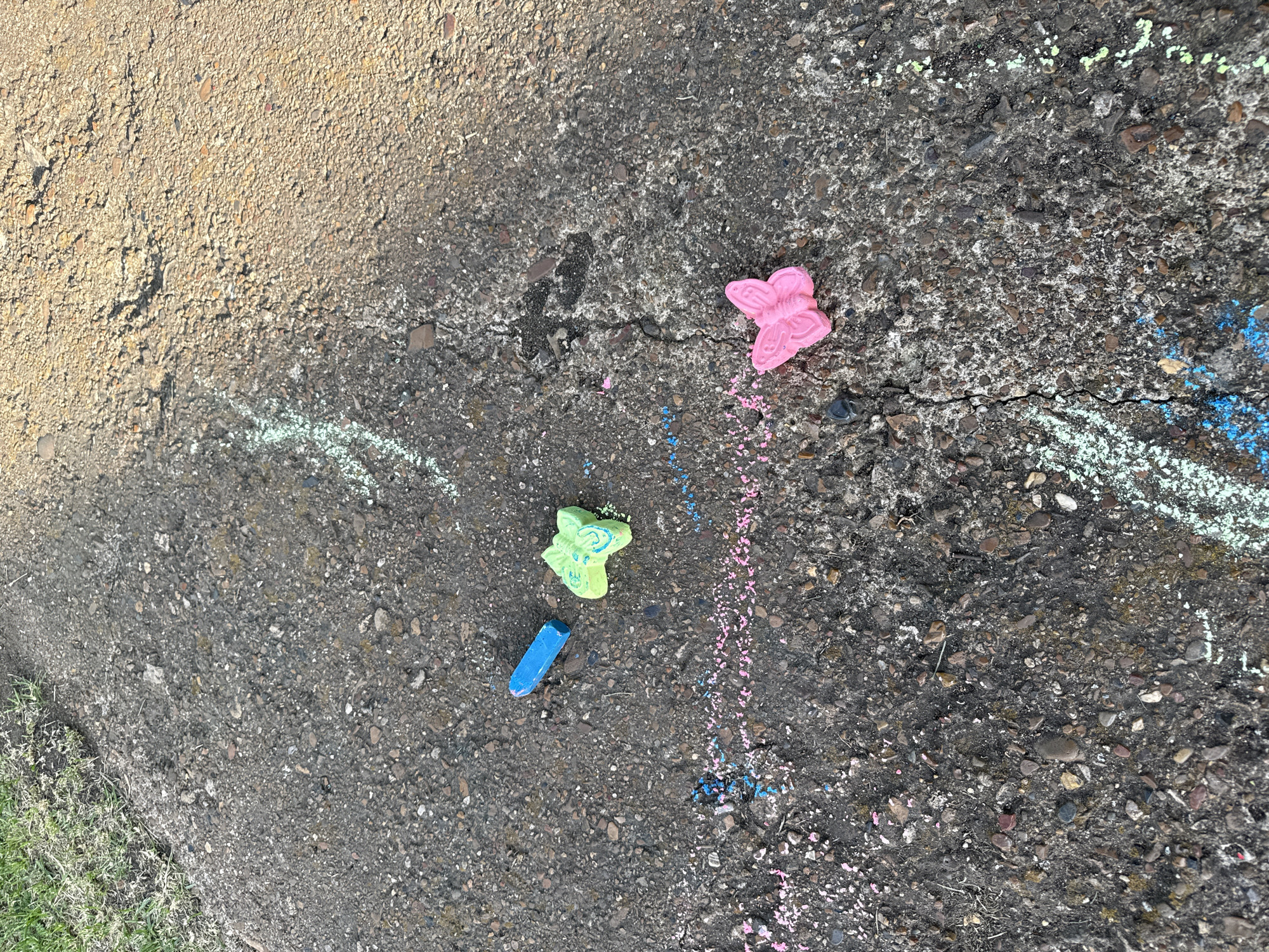 The image depicts a section of asphalt pavement with scattered chalk drawings and chalk pieces. A single blue chalk stick lies on the ground. A green chalk drawing in the shape of a butterfly is present, accompanied by some pink and blue chalk dust. Further along the pavement, a pink chalk butterfly is visible. Streaks of white, blue, and pink chalk mark the asphalt in various places. The pavement shows cracks and is a mix of dark gray and lighter brown tones. To the left of the pavement, a strip of green grass is visible, indicating an outdoor urban environment. The overall scene suggests recent children's activity in a public space.