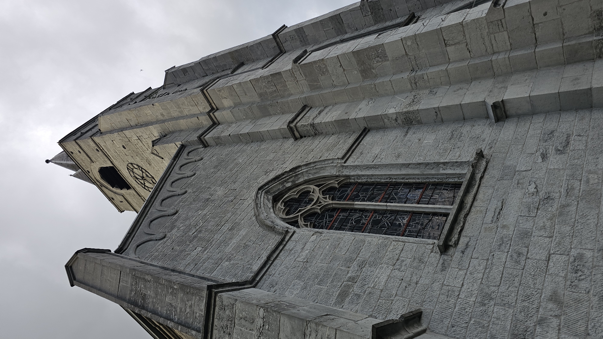 The image displays a low-angle perspective of a grand, historic stone building, likely a church or cathedral, situated in Boussu-lez-Walcourt, Belgium. The structure is constructed from large, grey, roughly hewn stone blocks, giving it a robust and ancient appearance.
In the mid-ground, a prominent Gothic-style window is visible. It features an arched top with intricate stone tracery, including a quatrefoil (four-lobed) design, and is filled with dark-paned glass, possibly stained glass. The window is set within a framed recess in the stone wall. Above and to the right of this window, decorative horizontal bands or cornices create a stepped, layered effect along the building's facade.
Further upwards and to the left, a taller section of the building's tower comes into view. This part of the tower displays a large, round opening, possibly a clock face or a rose window, with visible spokes or divisions. A small, pointed spire or pinnacle extends from the very top of this section.
The sky is overcast and uniformly grey, indicating a cloudy day and providing soft, diffuse lighting that highlights the textures and architectural details of the stone. There are no people visible, and no specific activity is taking place. The overall impression is one of majestic, enduring architecture. No discernible text is visible on the building.