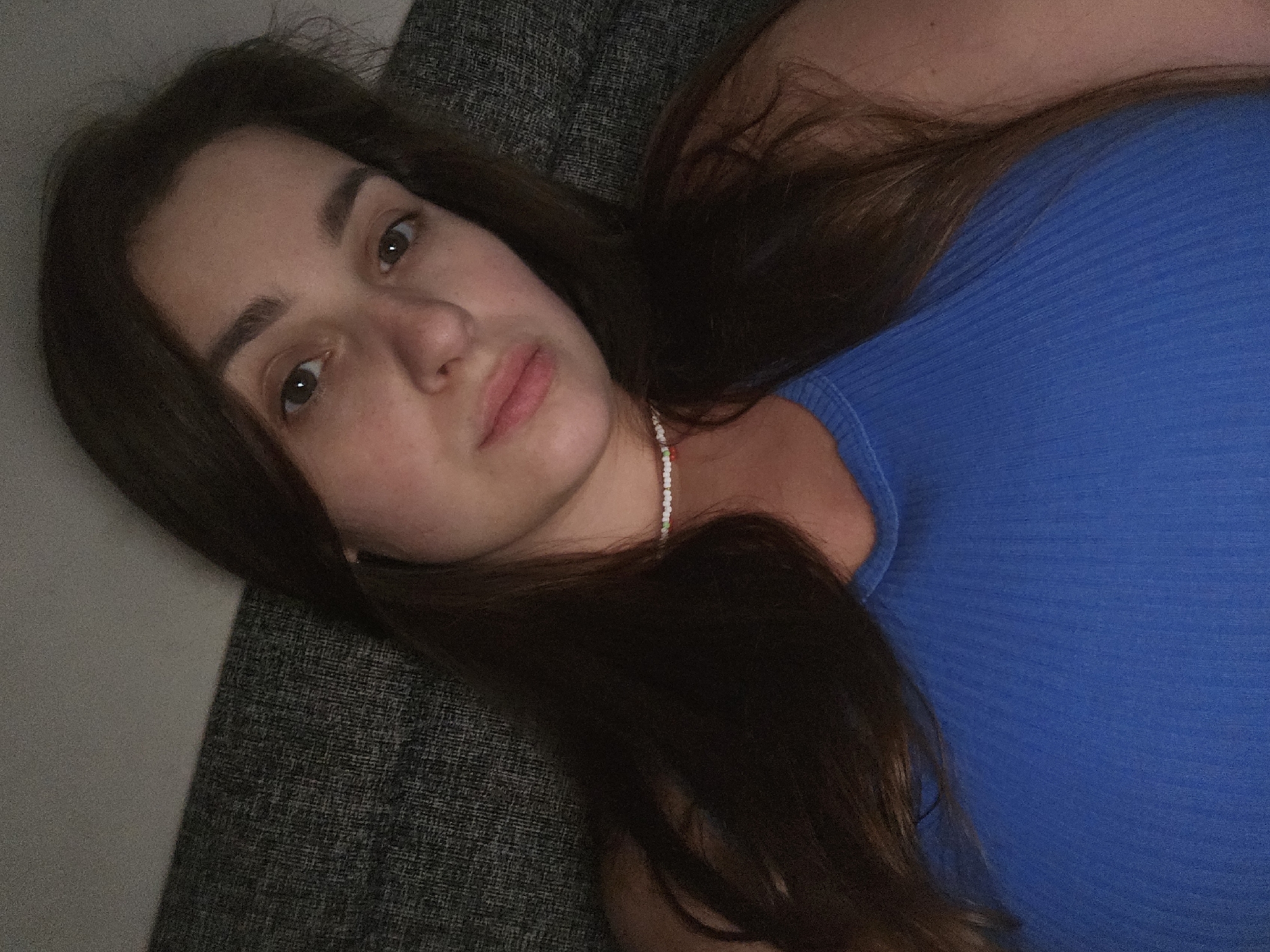 A selfie taken from a high angle shows a young woman with long, dark brown hair lying on a dark grey textured sofa. She is wearing a blue ribbed tank top and a beaded necklace. The lighting is dim, creating shadows on her face. The background consists of the sofa and a light grey wall. The scene appears to be an indoor domestic setting, likely a living room. There is no indication of the location being Petropavlivska Borshchahivka, Ukraine.