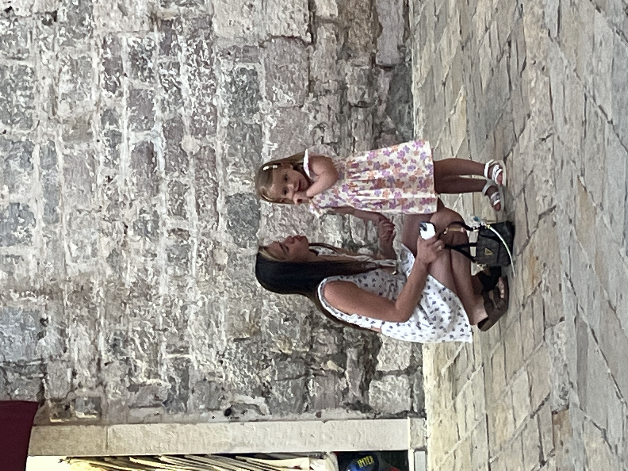 A woman in a white floral sundress squats down and holds hands with a young girl, who wears a similar floral-patterned dress. They are standing on a cobblestone street in front of a rough-hewn stone wall. The woman has dark hair and is wearing brown sandals. The girl has blonde hair in pigtails and is wearing white sandals. A small black handbag with a gold accent sits on the ground near the woman's feet. The scene appears to be outdoors, possibly in a historic part of the city of Budva, Montenegro, given the architecture.