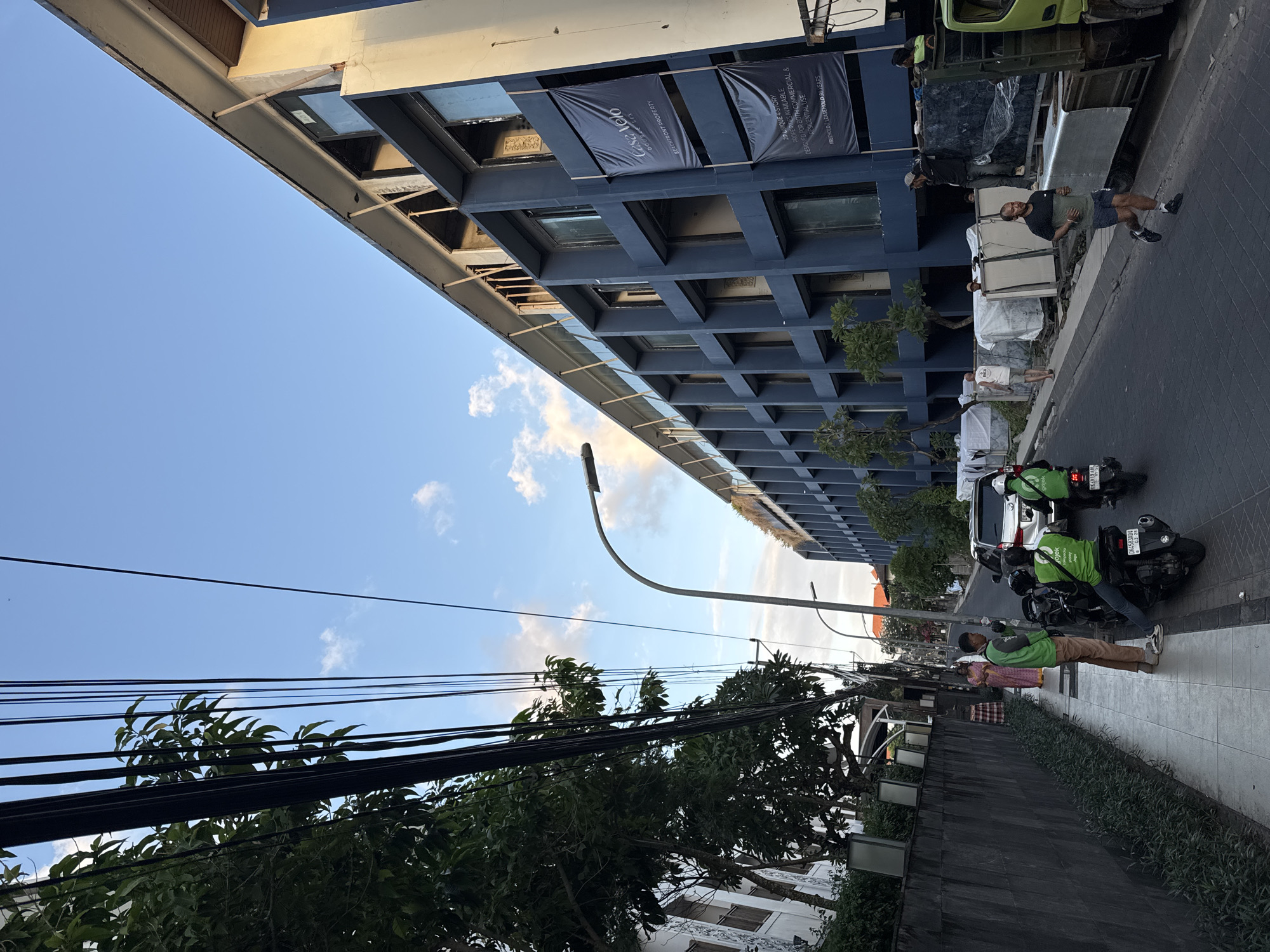The image depicts a street scene in Kuta, Indonesia, characterized by a multi-story blue building under construction or renovation on the right side of the frame. Overhead power lines and trees line the left side of the street, which is paved with dark cobblestones. Several individuals are present in the scene. On the left sidewalk, two men in green Gojek uniforms are standing near a parked motorcycle. Further down the sidewalk, a woman in a pink and gold outfit is walking. In the street, two Gojek motorcycles are moving, and a man is jogging. A white SUV is also visible in the traffic. Construction materials, covered in white tarps, are staged near a light green truck on the right side of the road. Two large black banners with white text are hanging from the blue building, one reading "Casa Velo DOUBLE SIX BEACHFRONT PROPERTY." The sky above is a clear blue with scattered white clouds, suggesting it is daytime, possibly late afternoon.