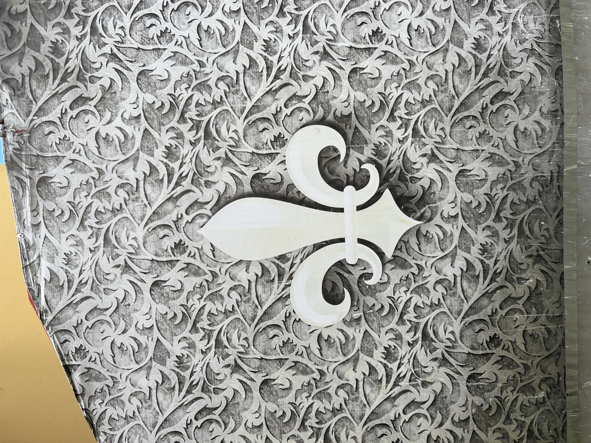 The image shows a close-up of a wall with a fleur-de-lis motif. The fleur-de-lis is a stylized lily or iris, and it is a symbol of royalty and French heritage. In this image, the fleur-de-lis is rendered in white and is overlaid on a patterned background. The background appears to be a wallpaper with a floral design in shades of gray and white. The overall impression is one of elegance and tradition. The image does not provide any information about its location in the city of Tomsk, Russia, or any details about people, actions, or interactions.