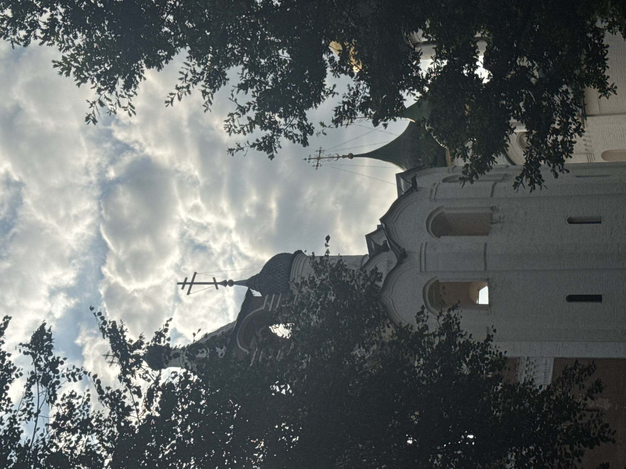 This image captures a view of the Suzdal Kremlin in Suzdal, Russia, with parts of the architecture partially obscured by dark green foliage. The sky above is filled with dramatic, textured clouds. Two onion domes, characteristic of Russian Orthodox churches, are visible; one is dark and topped with a cross, while another appears to be green and has a more ornate, gold-colored cross. A white, multi-story structure with arched windows is prominently featured in the foreground, suggesting it is a significant part of the complex, possibly a bell tower or part of a church. The lighting suggests it is either early morning or late afternoon.
