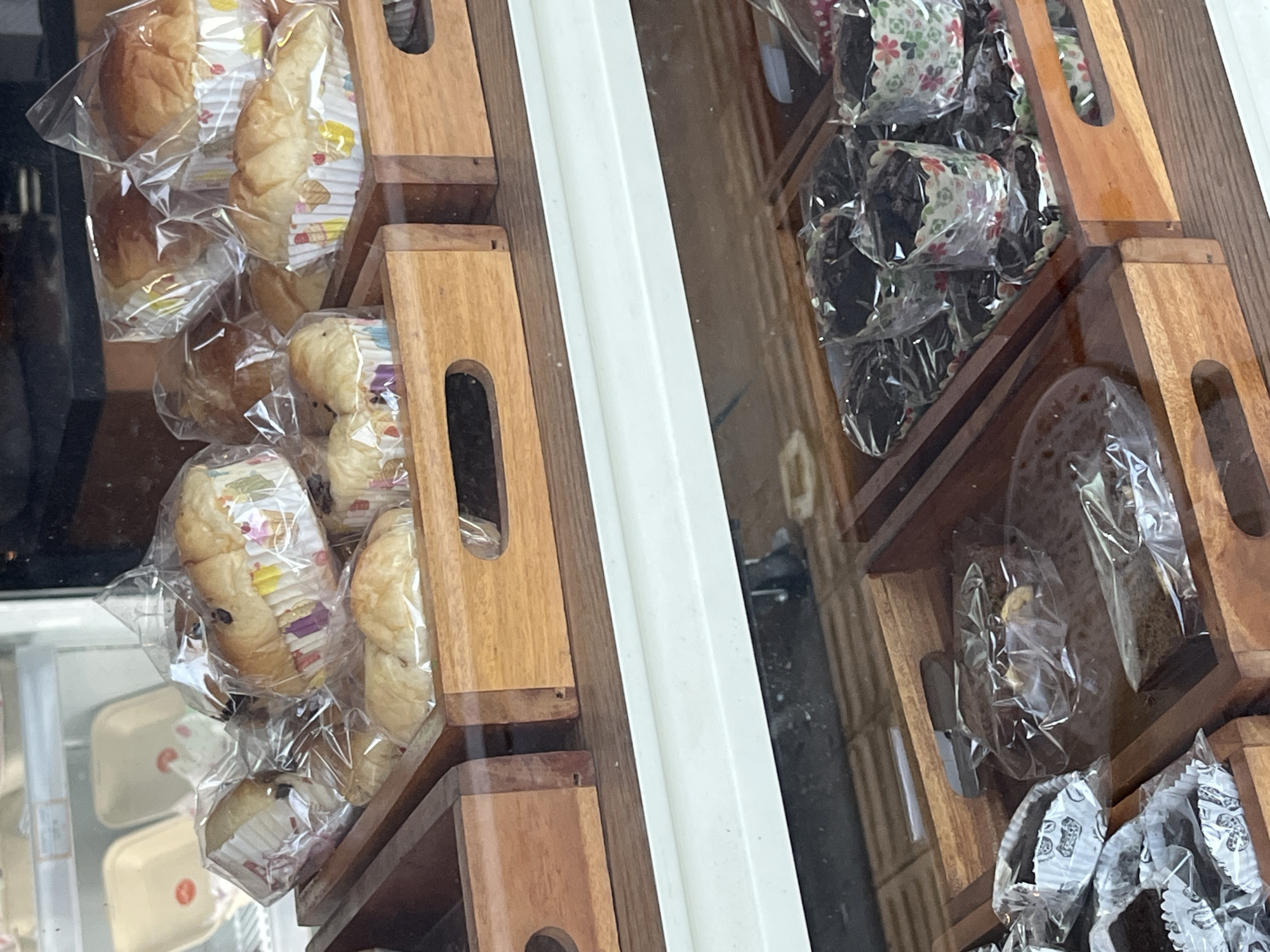 The image shows a display case of baked goods in what appears to be a bakery in Unknown, Indonesia. The baked goods are individually wrapped in clear plastic and arranged in wooden trays. The left side of the display features rolls and buns in decorative paper liners. The right side contains what appear to be chocolate cakes or brownies, also in paper liners. The display case is visible behind the baked goods. The scene is well-lit, suggesting an indoor environment.