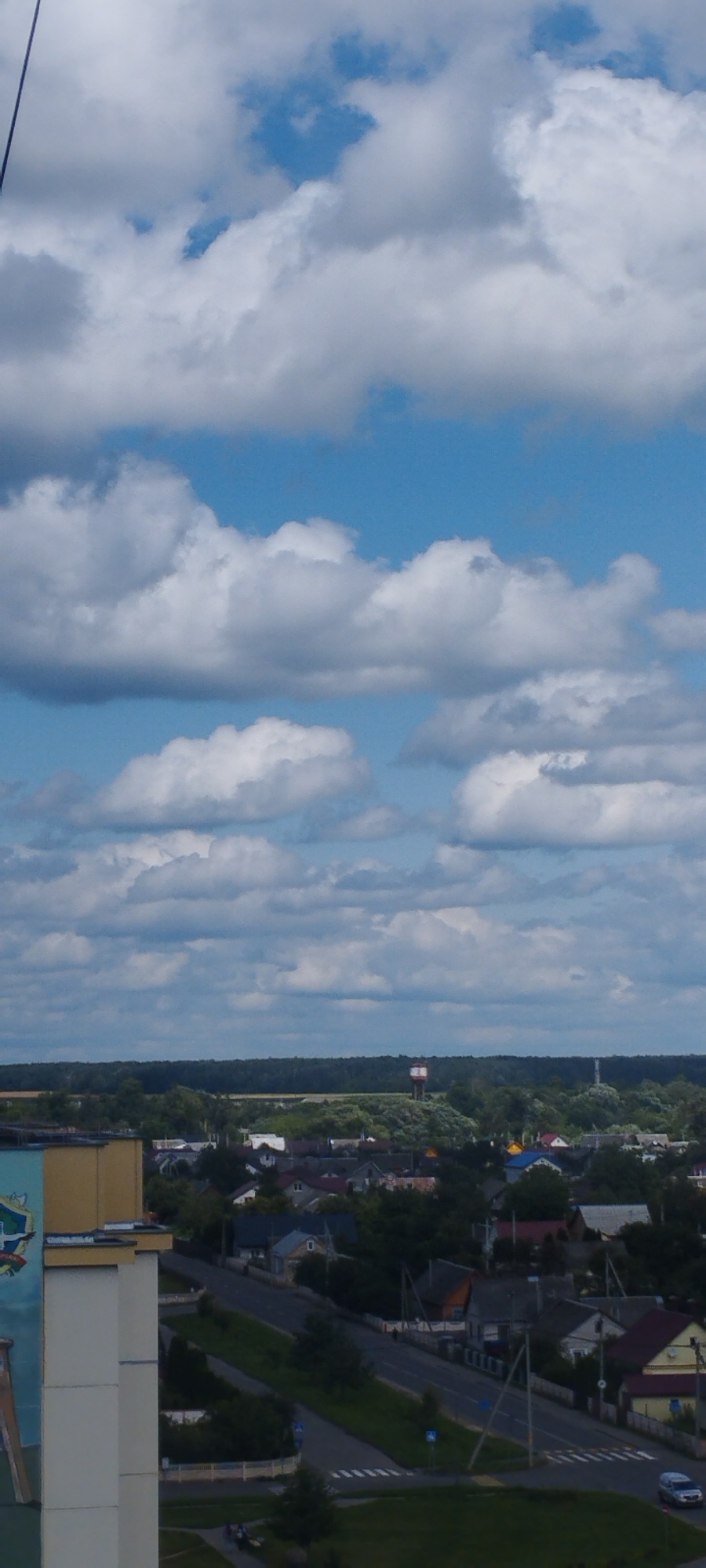 A vertical, eye-level, outdoor, wide shot shows a cloudy blue sky above a landscape of trees, buildings, and a road. The top of the frame is filled with white and gray clouds, some of which are bright and fluffy, while others are darker and more diffused. The sky is a bright blue, with patches of clear sky visible between the clouds.
Below the sky is a distant line of dark green trees, forming a dense forest that stretches across the horizon. In front of the forest, a small town or village is visible, with numerous buildings of various sizes and colors. The roofs of the houses are predominantly red, gray, or brown, and some have brightly colored accents. A prominent red and white water tower stands on a hill to the right of the forest.
A paved road winds through the village, with some cars and pedestrians visible. On the left side of the frame, the corner of a tall, modern building is visible, featuring a colorful mural on its side. The building appears to be made of white and yellow materials. The overall impression is that of a quiet, suburban or rural setting on a partly cloudy day.