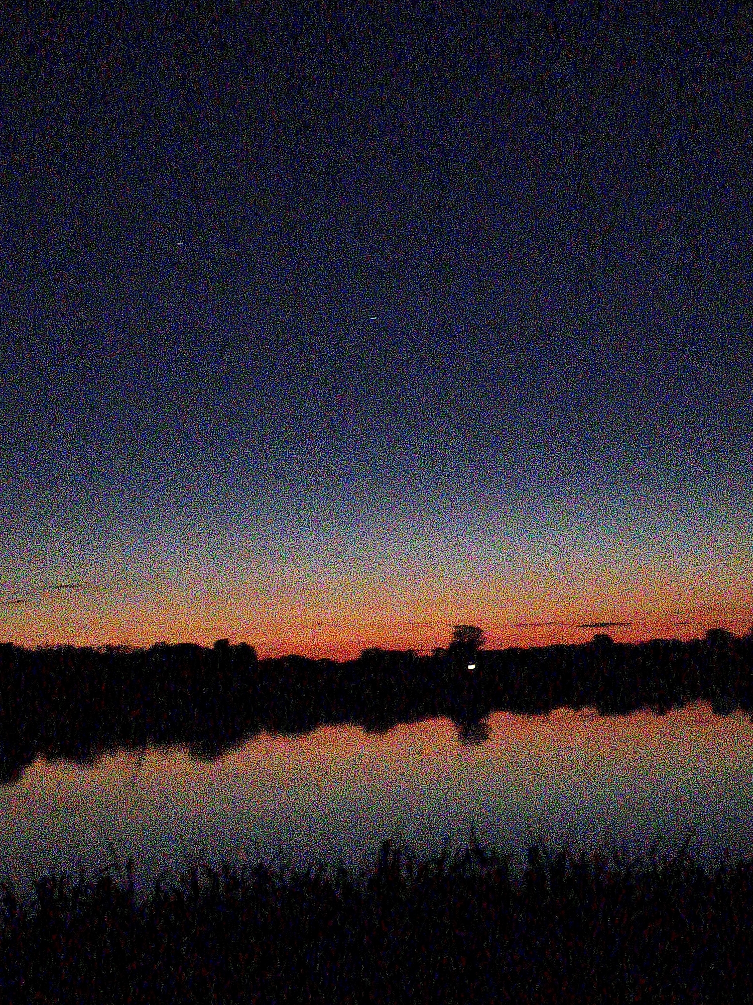 The image depicts a heavily pixelated and digitally noisy twilight landscape. The scene features a clear sky that transitions in color from a deep, dark blue-purple at the top to warm orange and red hues near the horizon, suggesting either dusk or dawn. A dark, irregular silhouette of what appears to be trees or a distant landmass spans horizontally across the middle of the frame, separating the sky from a body of water below.
The water surface acts as a mirror, reflecting the sky's gradient and the dark horizon line. The foreground is very dark, likely consisting of dense vegetation or ground cover. No people are visible.
Notable details include several small, faint white dots scattered across the darker upper sky, which could be stars or distant light sources. Near the center of the silhouetted horizon, a distinct, brighter vertical streak of white or light yellow stands out, possibly a distant artificial light, the moon, or another celestial body. Its reflection is clearly visible in the water directly below it. The overall impression is one of natural tranquility and stillness. There is no visible text in the image.
