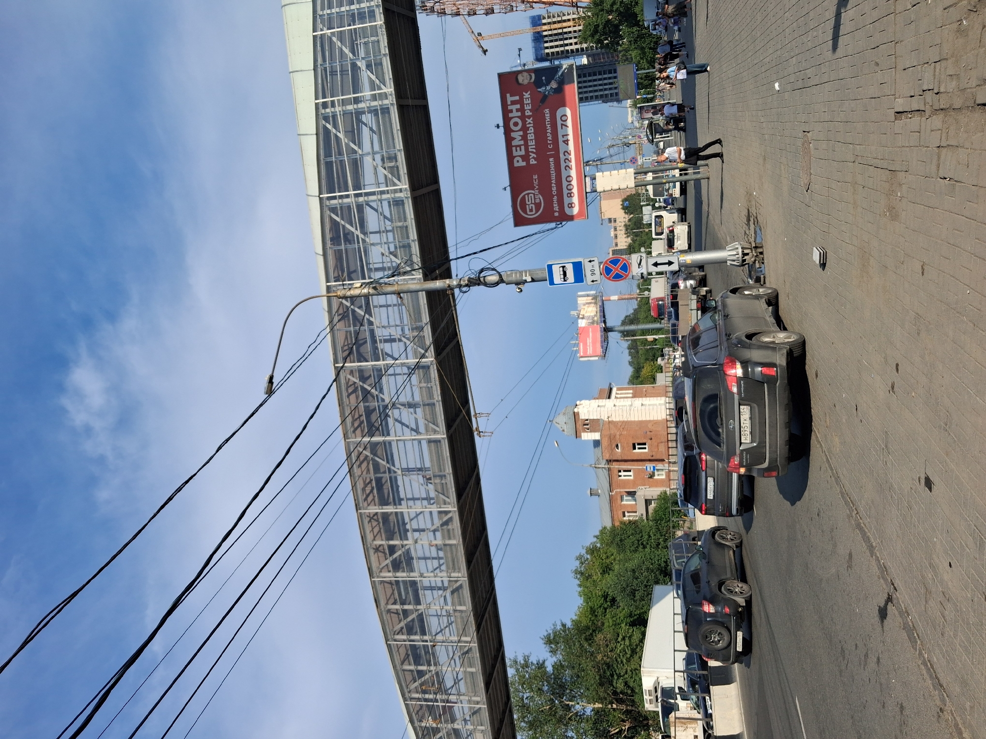 The image depicts a street scene in Novosibirsk, Russia, likely near an overpass. Multiple dark-colored SUVs are visible on the road, with one in the foreground showing its rear license plate H895TK 154. A pedestrian overpass, constructed of metal and glass, spans the road. A large red advertisement sign with text in Russian, including "РЕМОНТ" (REPAIR) and a phone number, is prominently displayed on the right side of the image. Traffic lights and street signs are present. Several people are walking on the sidewalk. A white truck is parked on the left side of the road. The sky is blue with scattered clouds.