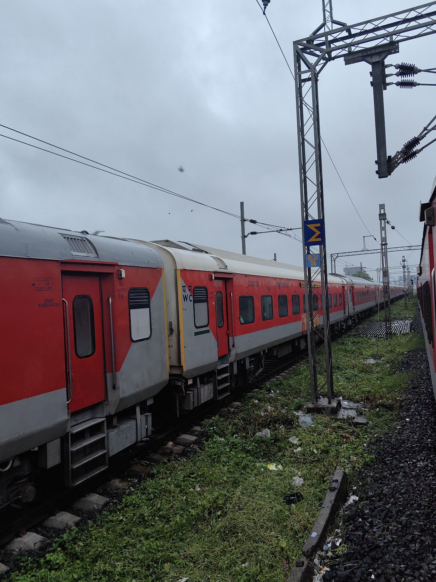 The image shows a red and grey train parked on a track next to a grassy embankment littered with debris. The sky is overcast and grey. A tall metal pole with overhead electrical wires stands to the right of the train. A blue sign with a yellow "M" symbol and text in Hindi is attached to the pole. The train appears to be at a station, as suggested by the platform and signage. The overall scene depicts a typical railway environment in India, likely on a cloudy day. Based on the distinct signage and train livery, this scene is identifiable as being in Bhopal, India.
