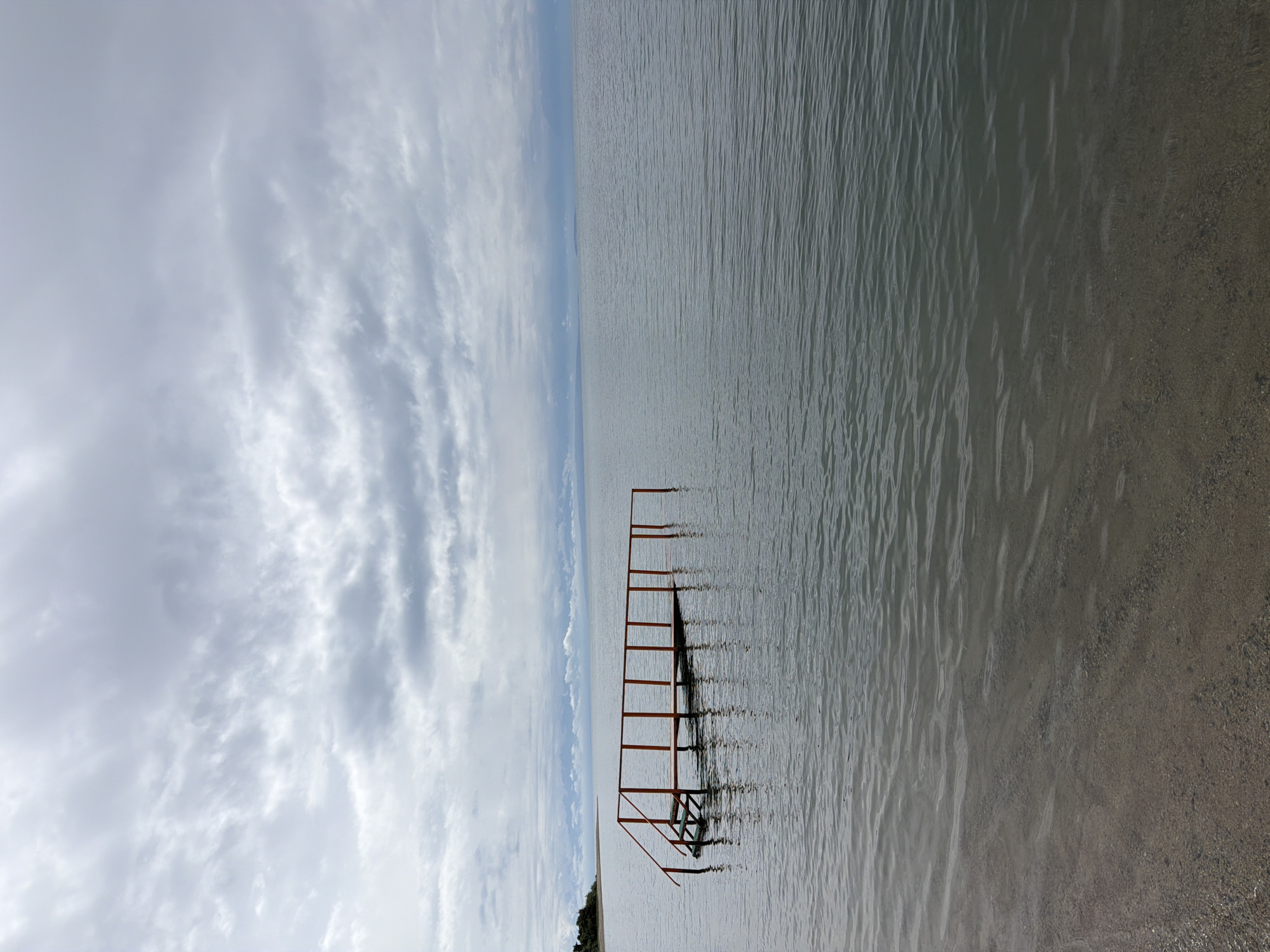 The image shows an outdoor scene with a partly submerged wooden pier in the foreground extending into calm water. The pier has red railings and steps. The water is a dark grey color and appears still with small ripples. In the background, there is a shoreline with some greenery and a wide expanse of water leading to a distant horizon. The sky is overcast with thick, grey clouds. The overall mood of the image is serene and perhaps a little somber due to the cloudy weather. The location is Unknown, Unknown.
