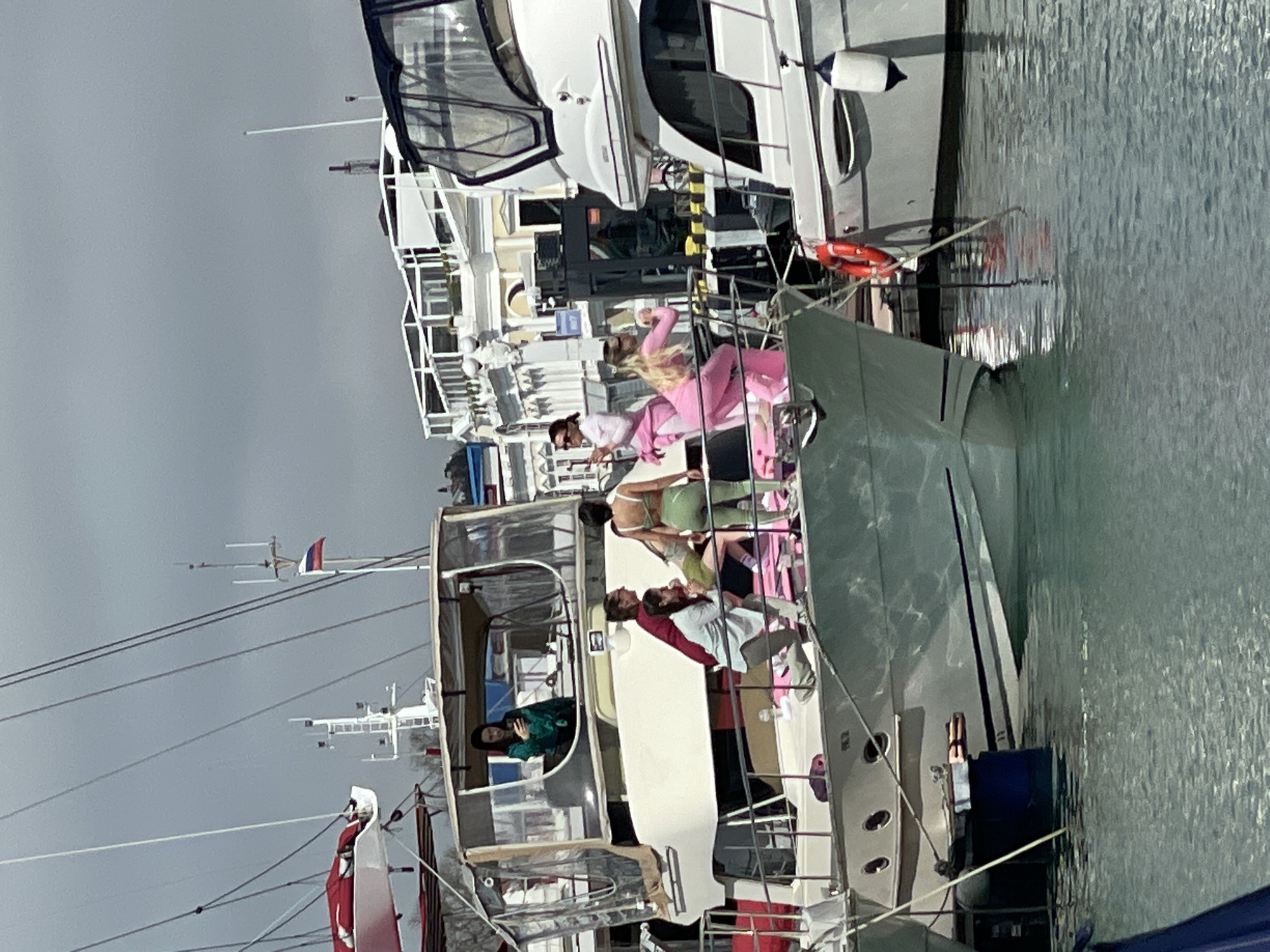 The image depicts a marina in Sochi, Russia, with multiple yachts docked. Several people are visible on the decks of the boats, some dressed in brightly colored clothing. The sky is overcast and the water is calm, reflecting the boats. There are power lines overhead. Flags of Russia are visible.