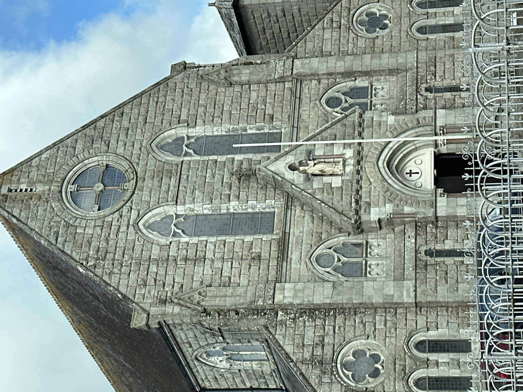 The image shows the exterior of a large, stone church in Ireland. The church has a traditional Gothic architectural style, with pointed arches, stained glass windows, and a large rose window at the front. A statue of a religious figure is visible above the entrance. The church is made of grey stone blocks. In front of the church is a white metal fence, behind which are parked several vehicles. The sky is partly cloudy.
