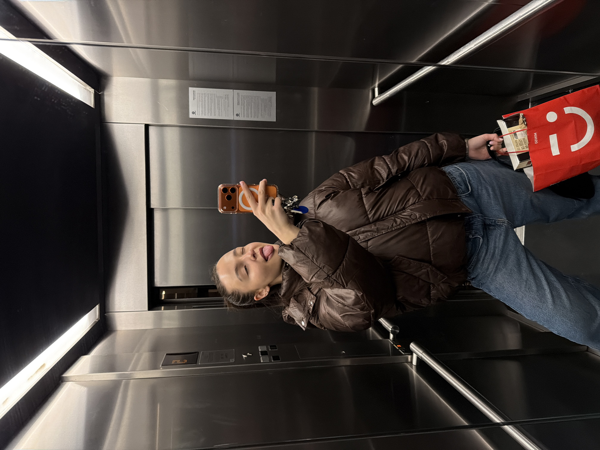 A young woman is taking a selfie in an elevator. She is wearing a brown puffer jacket and blue jeans, and she is sticking her tongue out at the camera. She is holding a phone in her left hand and a red shopping bag in her right hand. The elevator is made of stainless steel and has fluorescent lighting. There is a notice on the wall. The location is potentially in the city of Sofia, Bulgaria but this cannot be definitively confirmed from the image alone.