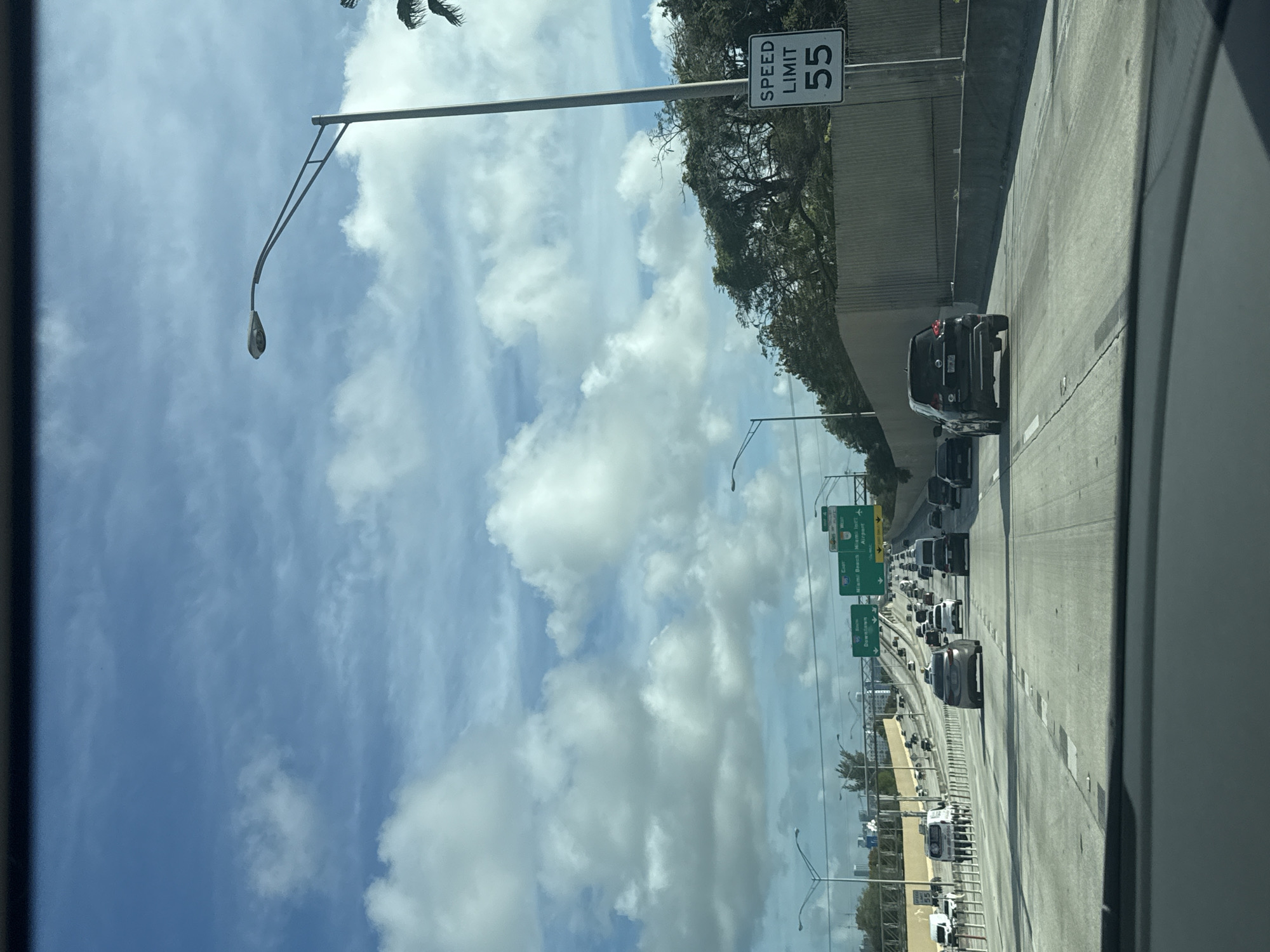 The image shows a highway scene in Miami, Florida, United States. The view is from inside a vehicle, looking out onto a multi-lane highway with moderate traffic. The sky is blue with scattered cumulus clouds. Highway signs indicate directions to "Downtown," "Miami Beach," "Miami Int'l Airport," and other locations. The speed limit sign reads "55." Vegetation borders the highway. Street lights are visible along the road.