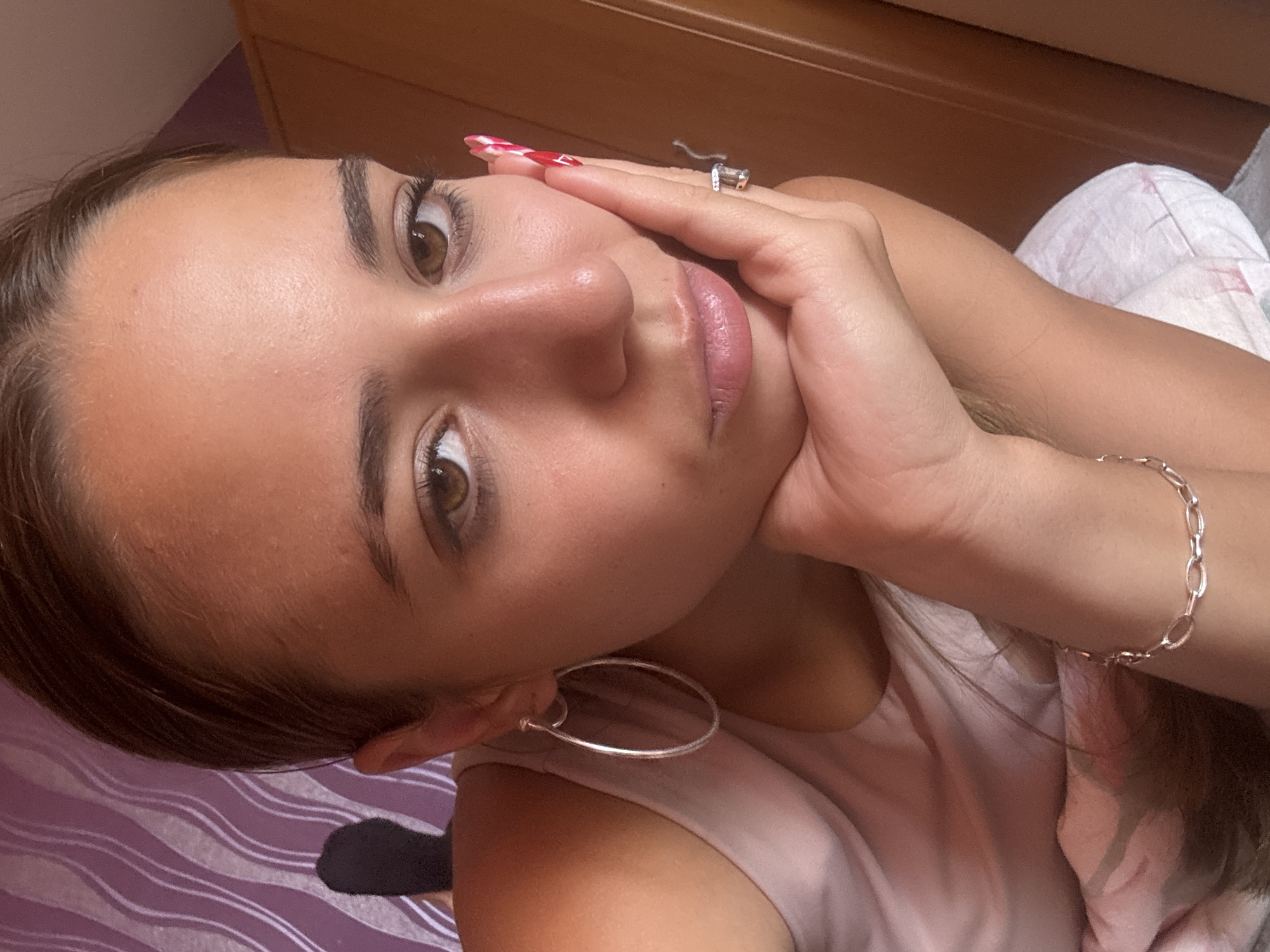 A young woman with brown eyes and brown hair pulled back poses for a selfie, resting her chin on her hand. She wears a light pink tank top, large silver hoop earrings, and a silver bracelet. Her right hand, which is supporting her chin, has bright red and white striped nail polish and a ring on her index finger. The background consists of a purple and white striped pattern on the left and a wooden wardrobe on the right. The scene appears to be indoors, likely in a bedroom, with natural lighting. There is no information indicating the location is Pernik, Bulgaria.