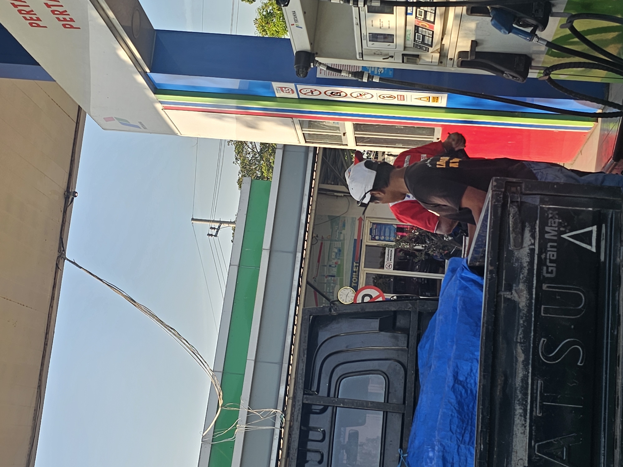 The image depicts a scene at a Pertamina gas station in Widang, Indonesia. In the foreground, a black pickup truck with a blue tarp covering its bed is parked. The tailgate of the truck displays the brand "DAI*TSU Gran Max". Two men are visible near a gas pump. One man, wearing a white cap and a black t-shirt with yellow lettering, is standing next to the pump. Another man in a red shirt is partially obscured behind him. The gas pump itself is white and blue with red and green accents, and has a Pertamina logo at the top. Behind the truck and the gas station structure, a building with a green roof and white siding is visible. The sky is clear and blue, suggesting it is daytime. Electrical wires hang from a pole in the background. A sign for "TOILET" is visible on the building.