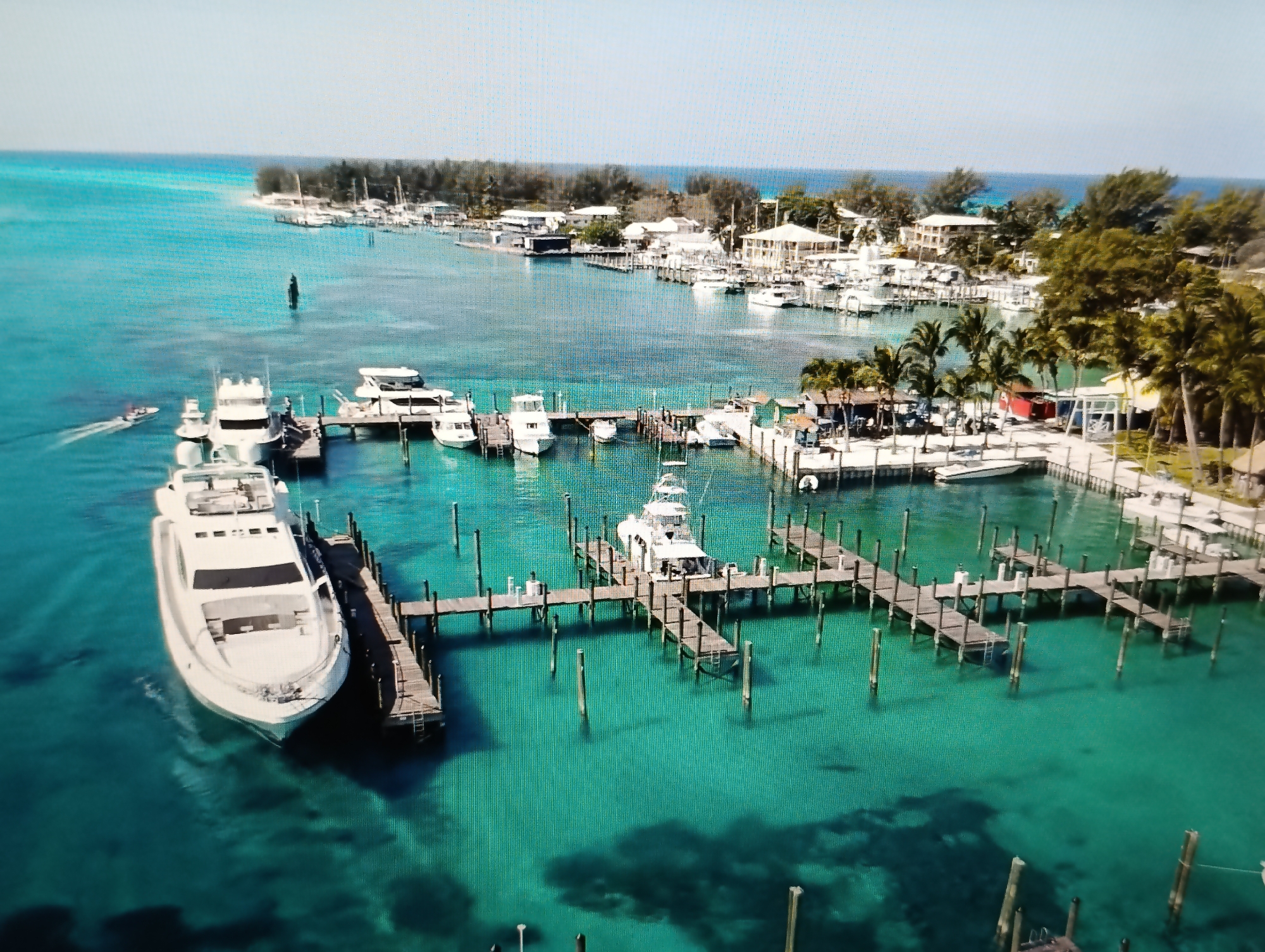 The image depicts a clear turquoise body of water with several boats docked at wooden piers. In the foreground, a large white yacht is prominently moored to a pier. Beyond this, more boats of varying sizes are seen at other piers, with some appearing to be fishing boats or smaller recreational vessels. The water is so clear that the seabed and shadows of the boats are visible beneath the surface. In the background, a coastline is lined with trees and buildings, suggesting a coastal town or resort. The sky is a pale blue, indicating a bright, sunny day. There are no visible people in the image. The location is identified as being in the city of Dubăsari town council, Moldova.