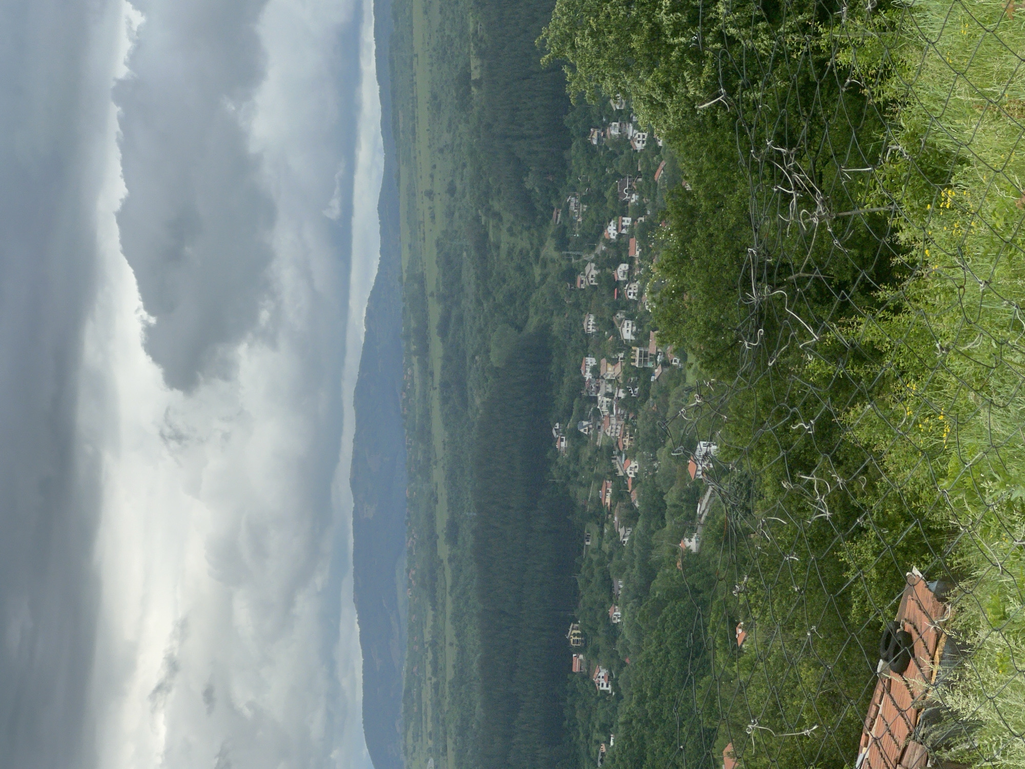 The image presents a panoramic view of a verdant valley and a village nestled on a hillside under an overcast sky.
In the immediate foreground, a black wire mesh safety net diagonally spans the lower right portion of the frame, with white ties visible on its knots. Green foliage, including grass, small yellow flowers, and tree leaves, grows through and around the netting. A small section of a reddish-brown tiled roof with two dark, round objects (possibly tires) on it is visible at the very bottom right, suggesting the photograph was taken from an elevated vantage point.
The midground showcases a village with numerous houses, characterized by white or light-colored walls and prominent red or brown tiled roofs, spread across a forested hillside. The village is surrounded by dense, green forests that cover the undulating terrain. Beyond the immediate village area, the landscape extends into a broad valley featuring lighter green fields and more scattered woodlands.
In the background, layers of hills and mountains recede into the distance, appearing somewhat muted in color due to atmospheric haze and the cloudy weather. The sky is dominated by a thick cover of grey and white clouds, indicating a cloudy daytime, with no direct sunlight visible.
The scene depicts a tranquil, rural landscape located in Marchaevo, Bulgaria. There are no people visible, and no specific activities are taking place. No text is visible in the image.