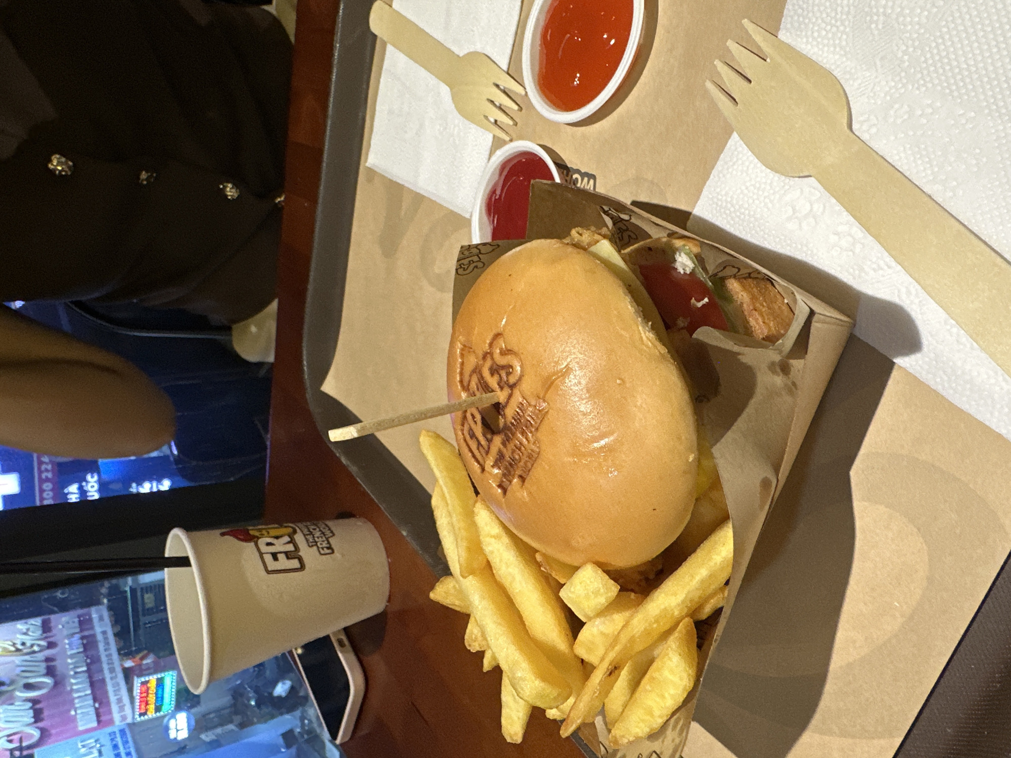 A high-angle shot shows a burger with fries, two cups of ketchup, a drink, and two wooden forks on a tray. Part of a person wearing a brown top with decorative buttons is visible on the left. The scene is likely in an eatery, with reflections of buildings and text visible on a digital screen below the tray. Text is "HA" and "UOC". The city is Thủ Đức, Vietnam.