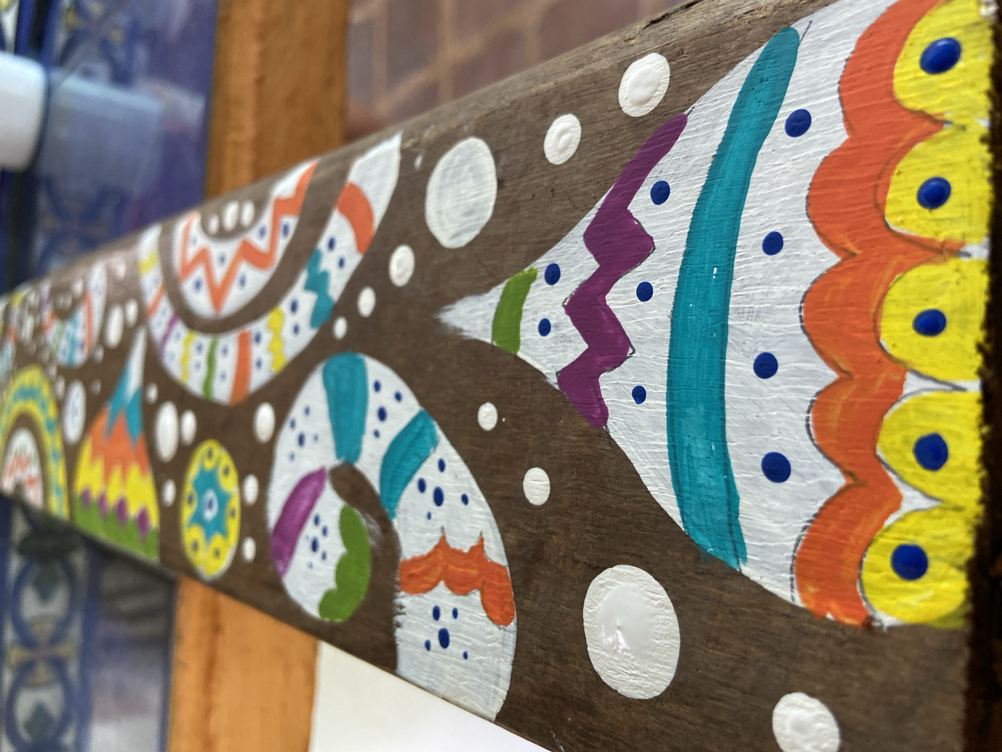 A wooden beam, angled diagonally, is the central subject of this close-up shot. The beam is adorned with a vibrant, hand-painted pattern that combines abstract and stylized motifs. The base of the painting is a solid white, onto which colorful shapes and designs are layered. Prominent colors include bright orange, yellow, various shades of blue, green, and purple, contrasting with the dark brown, textured wood of the beam.
The patterns feature zigzags, wavy lines, and numerous dots of different sizes and colors. Small blue dots are scattered within larger painted shapes, while larger, raised white dots are interspersed across the unpainted wooden sections, adding a tactile texture to the surface. On the far right, a sequence of orange and yellow scalloped shapes, each containing a raised blue dot, creates a striking border or repetitive motif. Further left, there are elements resembling stylized feathers or leaves, alongside concentric circles and triangular designs in different hues. The overall style suggests a folkloric or indigenous artistic influence, possibly reflecting the local culture of Los Velásquez, Venezuela.
The background is out of focus, providing depth without distracting from the painted beam. On the left, blurred blue surfaces with faint circular patterns are visible, possibly tiles. To the right, a soft, blurred reddish-brown surface can be seen, which might be another wooden structure or a wall. The lighting suggests daytime, with soft illumination highlighting the textures of the paint and wood. There is no visible text in the image.