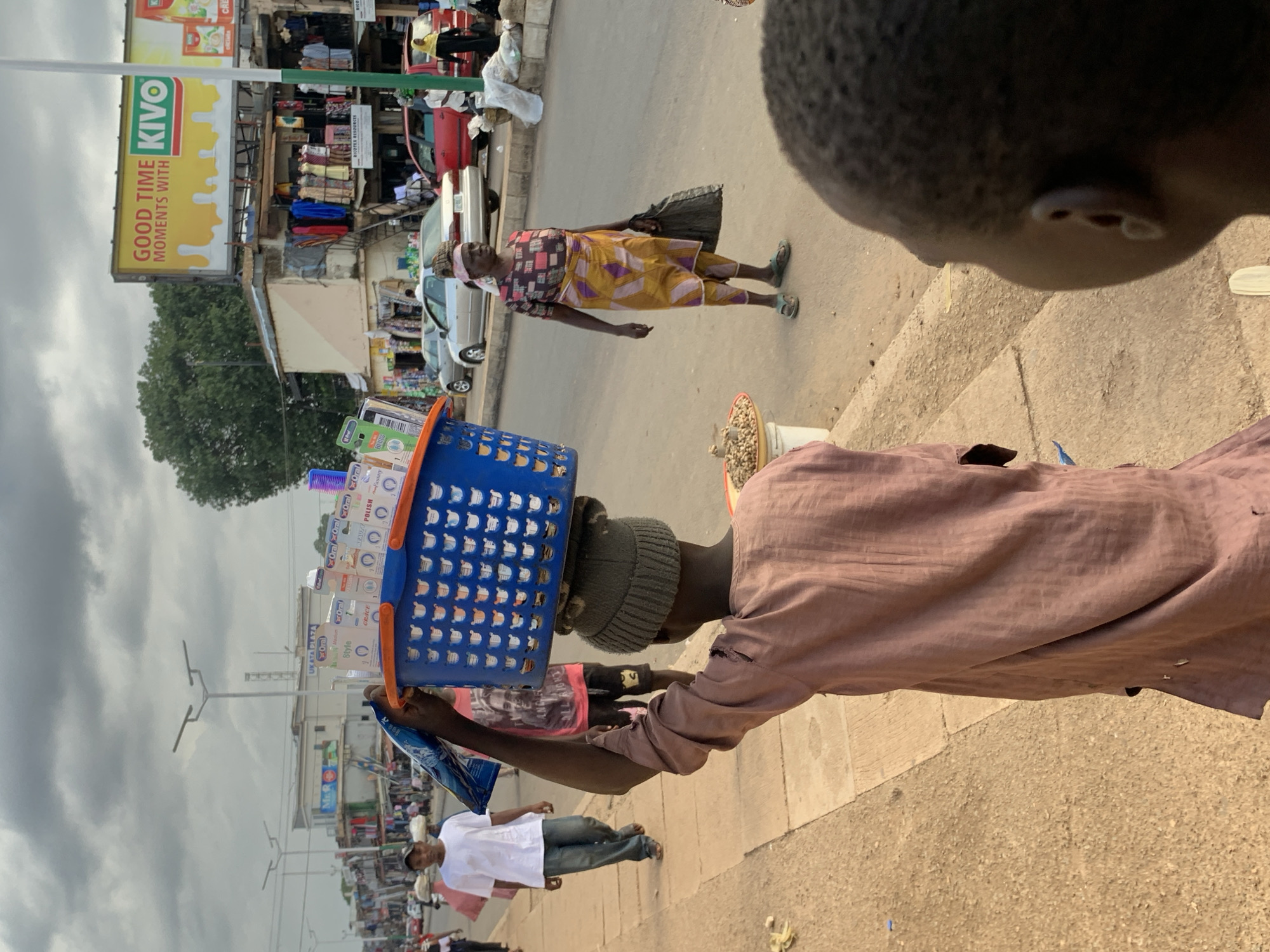 The image depicts a street scene in Jos, Nigeria, on a cloudy day. In the foreground, a young person wearing a pinkish-brown tattered robe and a grey beanie carries a blue basket filled with oral care products on their head. To the right, a woman in a colorful patterned outfit and a yellow wrap skirt stands with a black bag in her hand. In the middle ground, a man in a white t-shirt and jeans walks away from the camera. The street is lined with businesses, including a store with clothing displayed outside, and a large billboard advertising "GOOD TIME MOMENTS WITH KIVO." Several cars are parked and driving on the road. The environment suggests a busy commercial area with pedestrians and vendors.