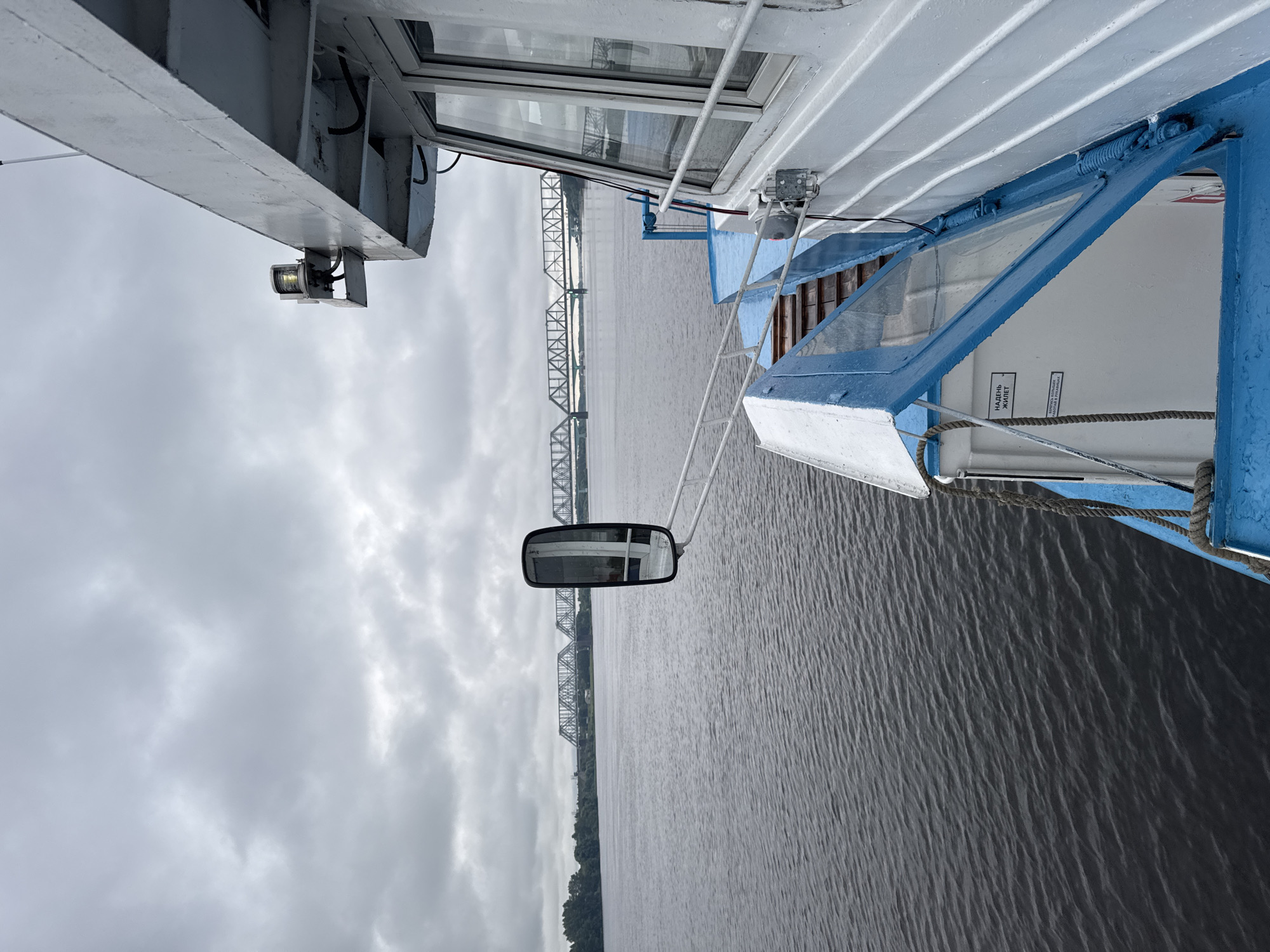 A side view of a white and blue boat navigates on dark, rippling water under a cloudy, overcast sky. The boat's deck is visible on the right, with a windowed cabin and an open hinged panel. A side mirror is mounted on a white arm, reflecting the cloudy sky. In the distance, a long, grey metal railway bridge spans across a wide body of water, with a treed shoreline visible on the left. The scene is devoid of any visible people. The image appears to be taken from within the city of Шебунино, Russia, given the context of the surrounding area.