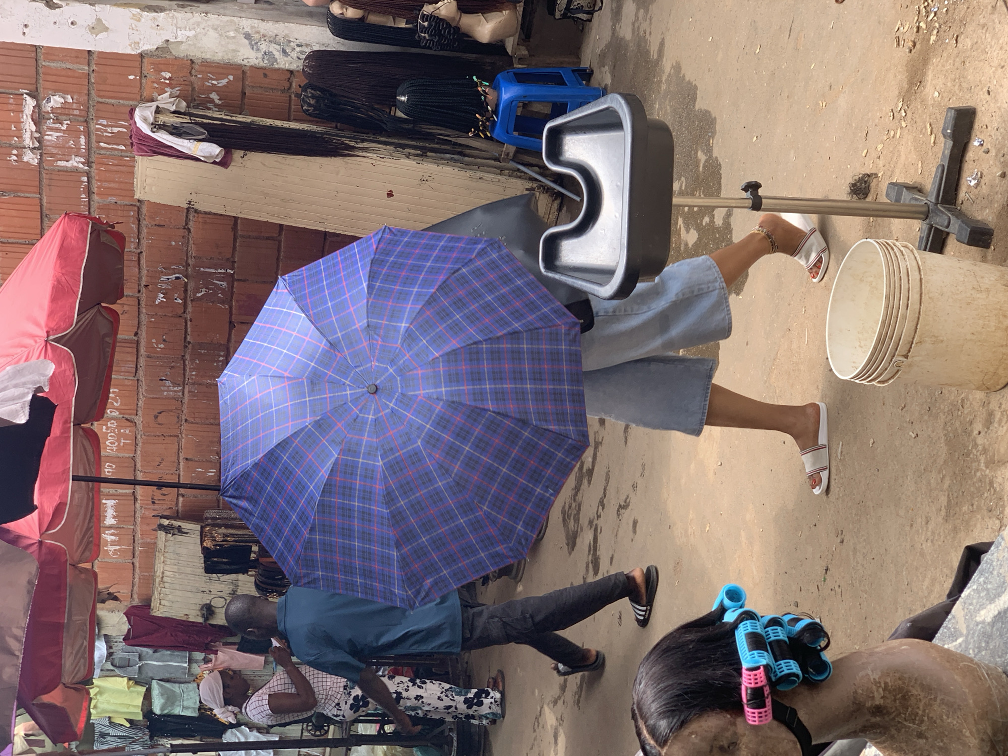 The image depicts an outdoor market scene in Abuja, Nigeria. In the foreground, a mannequin head with hair rollers is visible. A person is walking past holding a large blue and purple plaid umbrella. To the left, two individuals are interacting under a red awning, with clothing and accessories displayed behind them. Further back, a brick wall serves as a backdrop, with various wigs and hair products displayed on shelves and stands. A portable hair washing station with a black basin and a metal stand is positioned in the middle ground, next to a white bucket. The ground is dusty and uneven, typical of an outdoor market environment. Several other individuals are partially visible in the background, suggesting a busy marketplace.