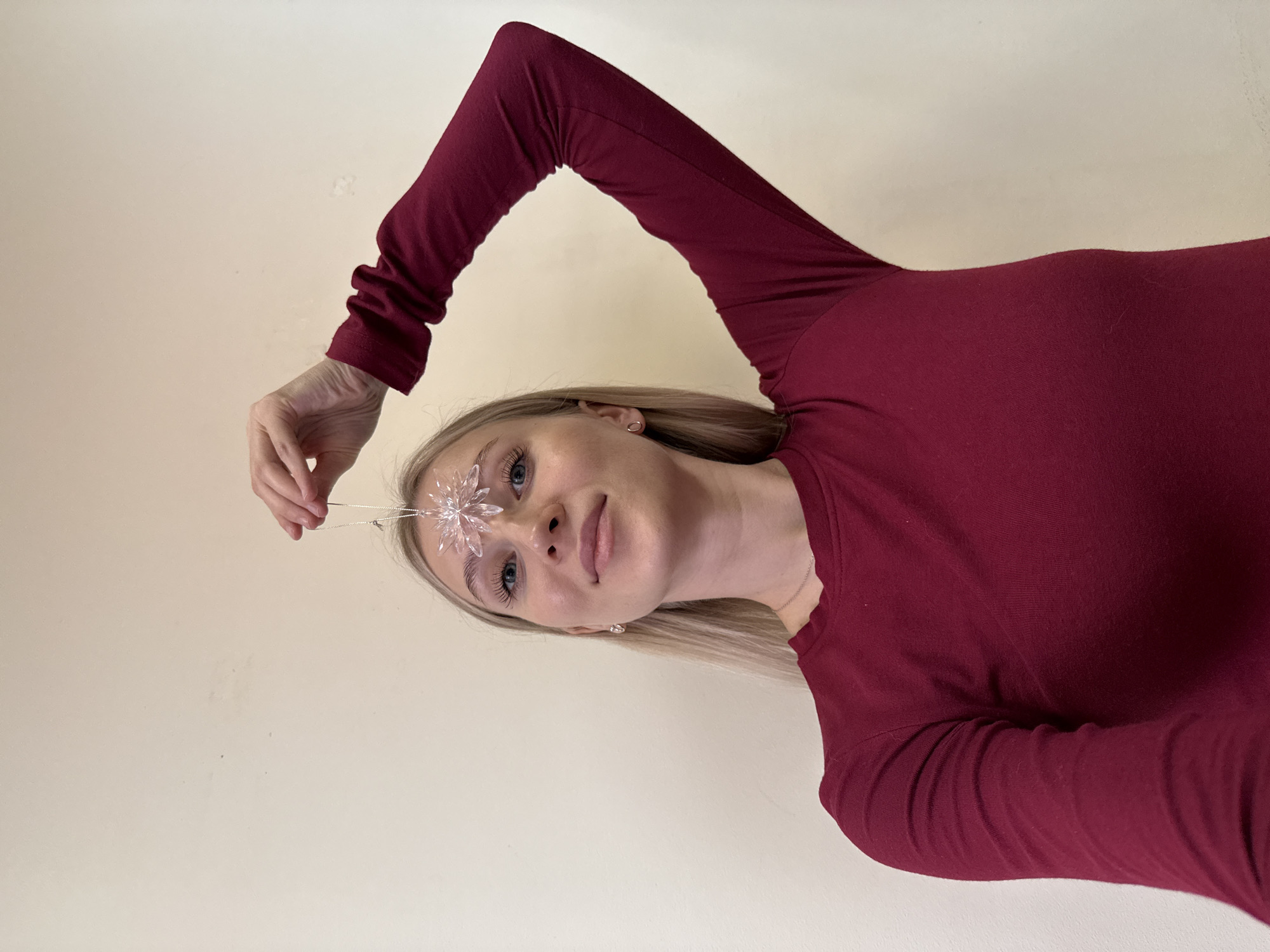 A woman with light blonde hair and blue eyes is holding a crystal snowflake decoration above her forehead. She is wearing a long-sleeved maroon shirt. The background is a plain, light-colored wall. The image is rotated, appearing as if the woman is looking down from above. There is no evidence in the image to suggest a location in Izhevsk, Russia.