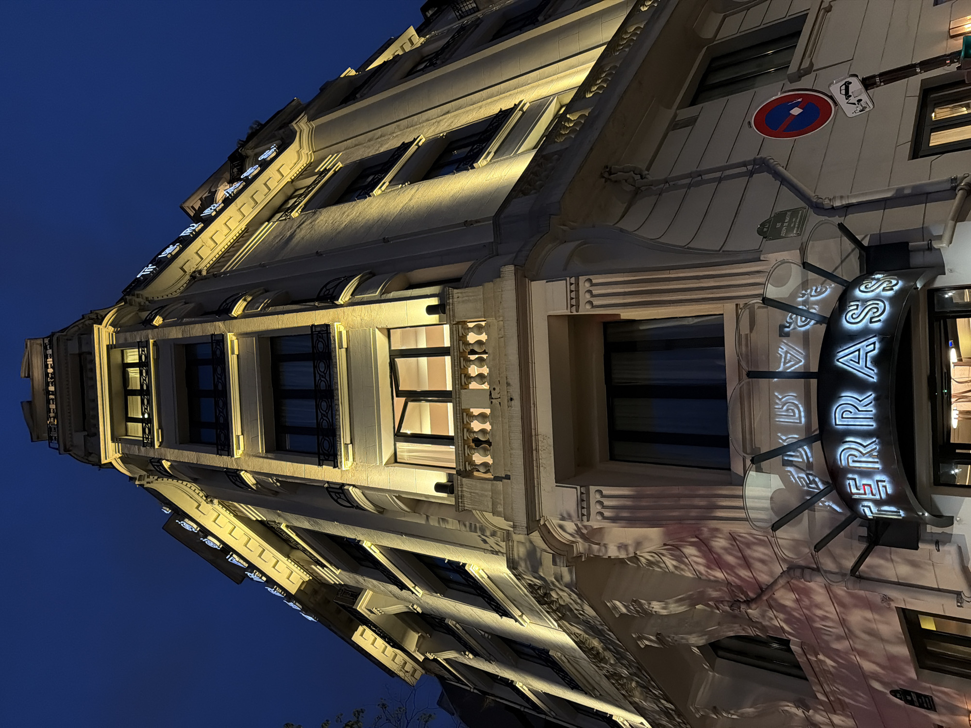 This is an upward-angled nighttime shot of a light-colored building in Paris, France, taken at an oblique angle. The building is illuminated with warm lighting accentuating its architectural details. "TERRASS" is written on a lighted sign next to an entrance. A round blue and red no-parking sign is visible near the top of the building, beside another sign for loading and unloading. The dark blue sky appears in the upper portion of the frame. The building has multiple windows with dark frames, some with decorative elements. The overall impression is of a classic Parisian architectural style at dusk or night.