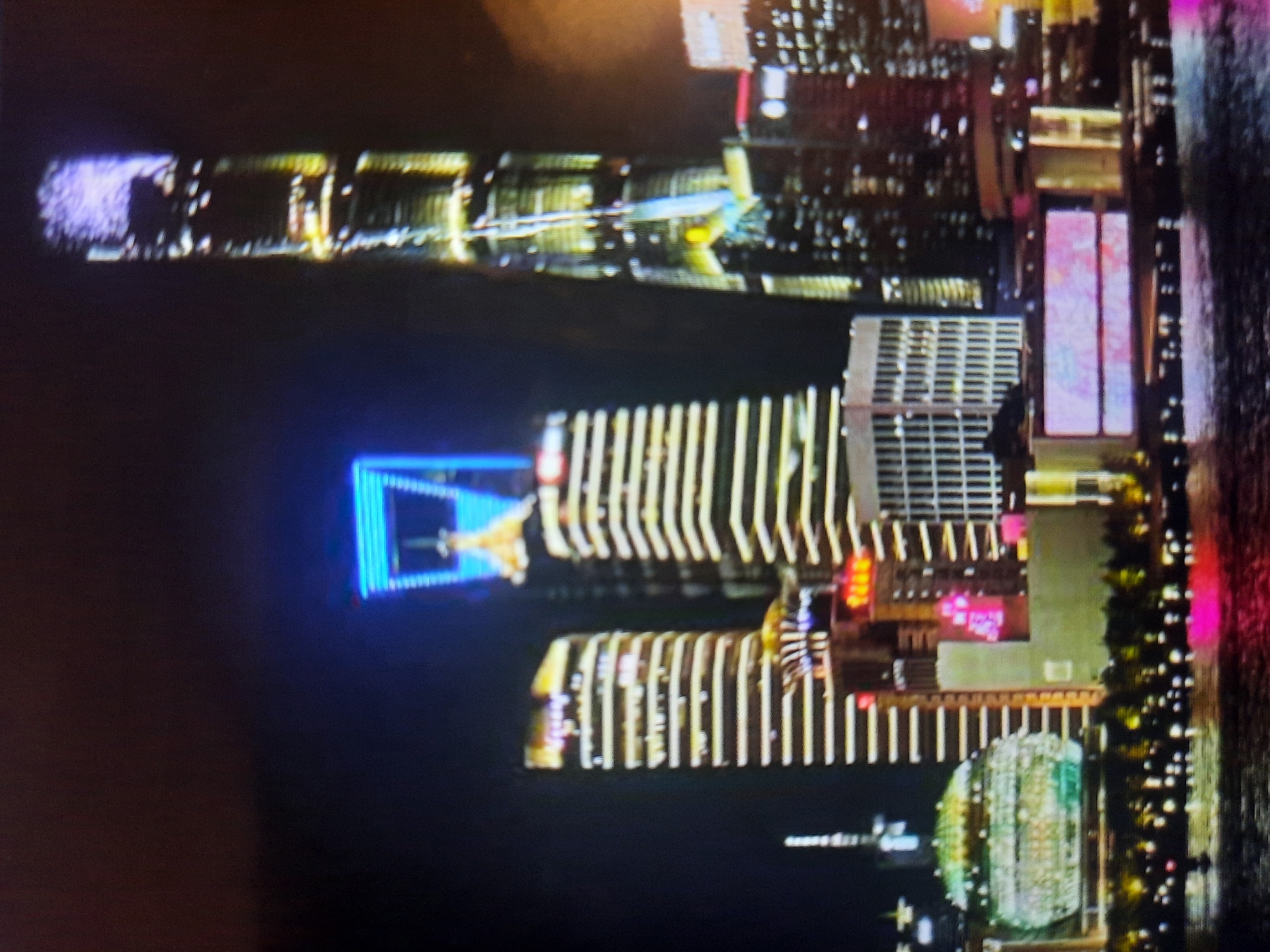 This image depicts a nighttime view of a modern city skyline, characterized by numerous illuminated skyscrapers. The buildings vary in height and architectural design, with some featuring horizontal banding of lights and others with more complex illuminated patterns. One exceptionally tall skyscraper in the center-right of the image is prominently lit with a vertical strip of lights and has a brightly illuminated, iridescent spire. To the left of this central skyscraper, another tall building displays a large, rectangular blue light outline near its apex.
Further to the left, a distinctly dome-shaped building with a patterned, glowing facade is visible, reflecting light onto the water in the foreground. The foreground consists of a body of water, likely a river or harbor, reflecting the city lights. The water surface is dark, with scattered points of light and a pinkish hue on the right side. The overall atmosphere is one of urban vibrancy and nighttime illumination. The city of Rosenheim, Germany is not depicted in this image.