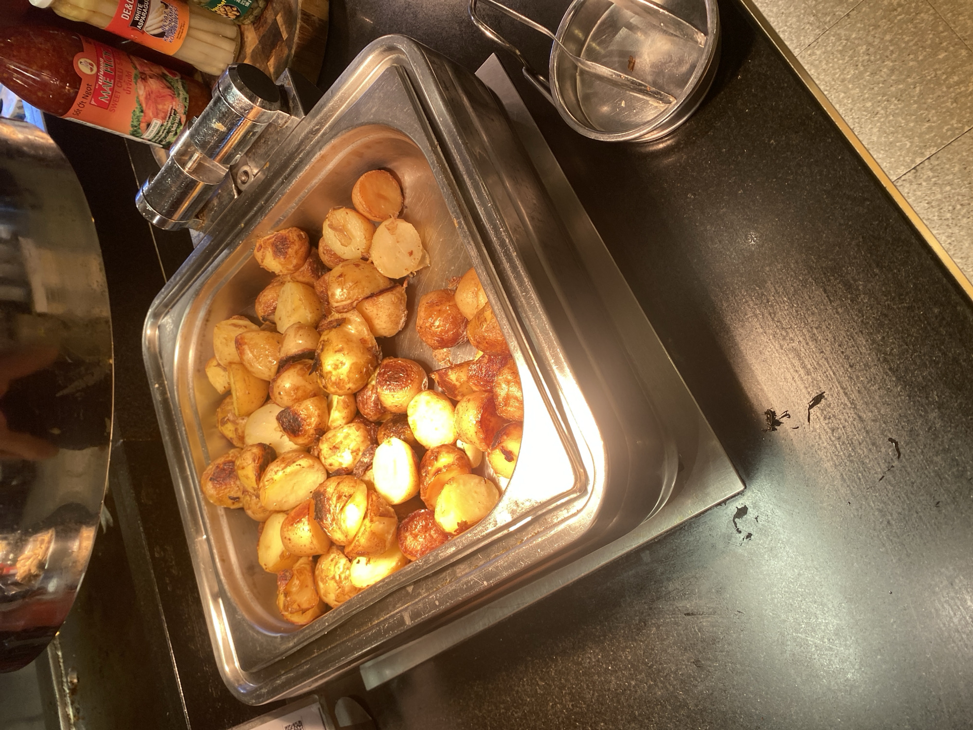 In Aksu, Turkey, a buffet-style display features a large rectangular stainless steel pan filled with roasted potatoes. The potatoes are small to medium in size, halved, and appear golden brown with some charred edges, suggesting they are seasoned and baked. The pan is set within a larger stainless steel warming unit, which rests on a dark countertop. To the right of the pan, a silver metal tong holder with tongs is visible. In the background, partially out of focus, are two bottles: one is a red chili sauce bottle, labeled "Mae Ploy Sweet Chili Sauce," and the other appears to be a jar of white asparagus. The overall environment suggests a food service setting, likely a restaurant or hotel buffet.