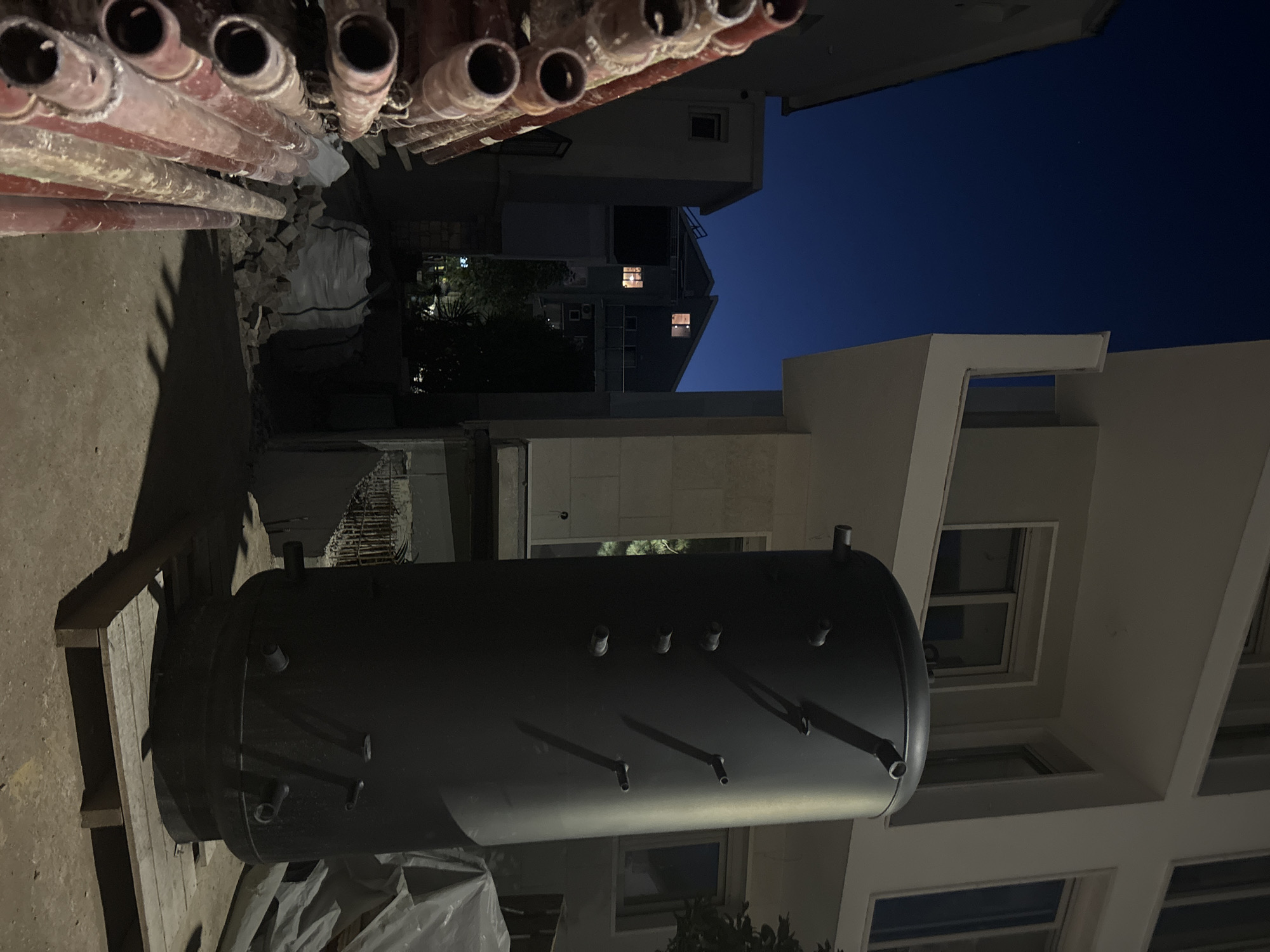 A black cylindrical water heater stands on a wooden pallet in front of a light-colored modern building. The scene is illuminated by artificial light and the fading twilight sky, which is a deep blue. To the left, a stack of red scaffolding pipes is visible, angled upwards. In the background, across from the modern building, are other residential buildings, some with lit windows. The ground is paved and appears to be part of a construction site, with rubble and a large white bag of material visible. The overall setting suggests a residential area under development in Budva, Montenegro, at dusk.
