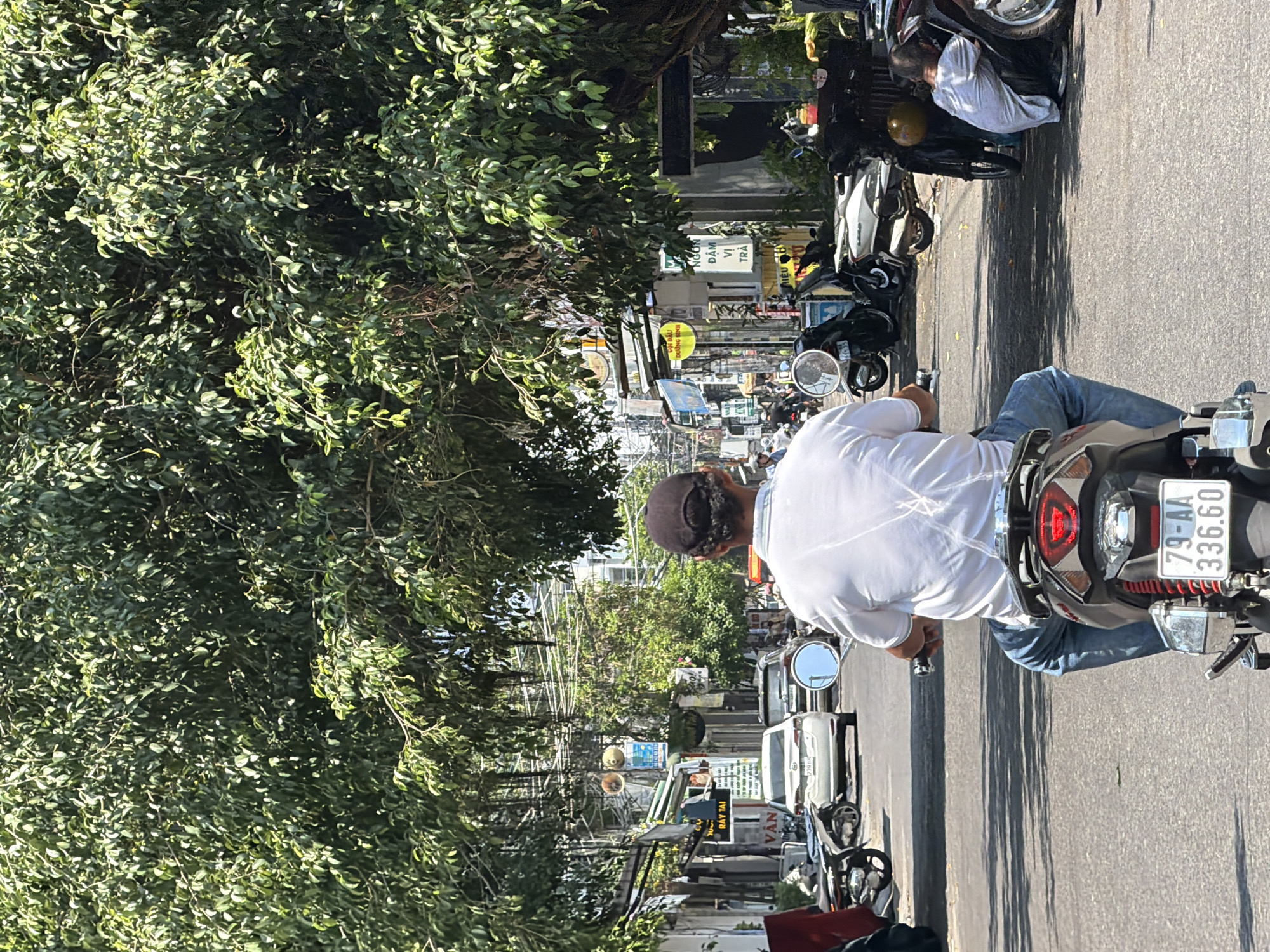 The image shows a street scene in Nam Nha Trang Ward, Vietnam. A person, seen from behind, is riding a scooter down a road lined with buildings and trees. The rider is wearing a white t-shirt, blue jeans, and a dark baseball cap. The scooter has a license plate that reads "79-AA 336.60". Other scooters and cars are parked on the side of the road, and there are signs and wires visible on the buildings. One person is seen squatting down near a parked scooter on the right side of the image. The trees are large and leafy, casting shadows on the road. The sky is visible above the trees.