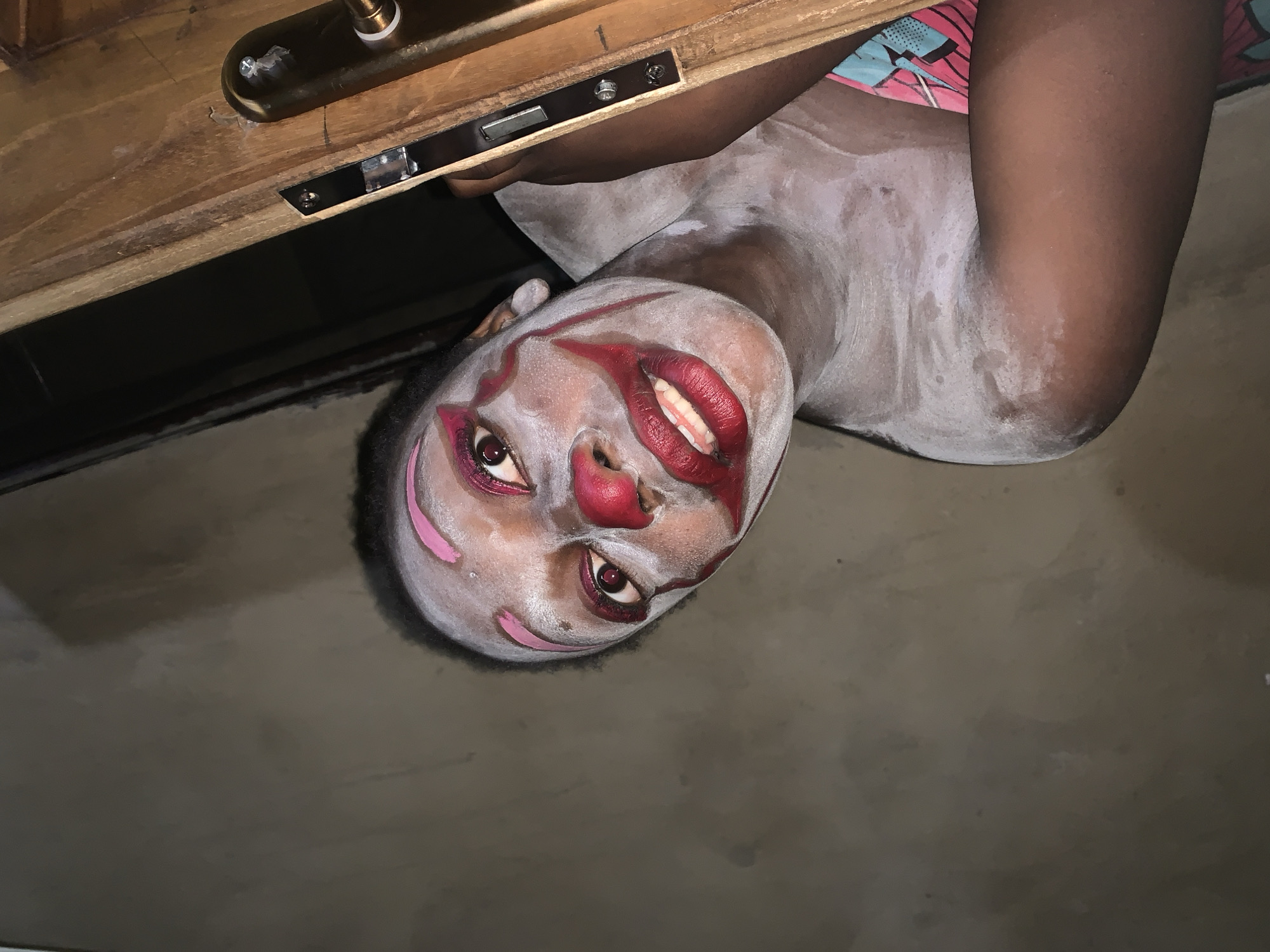 A person with clown makeup is peeking through a doorway. The makeup includes white base, pink eyebrows, red cheeks, and a red clown nose. The person's lips are painted red. The person is wearing a colorful top. The scene appears to be indoors, possibly in a home, given the wooden door and what looks like a wall in the background. The lighting is dim. The location is stated to be Agban, Nigeria.