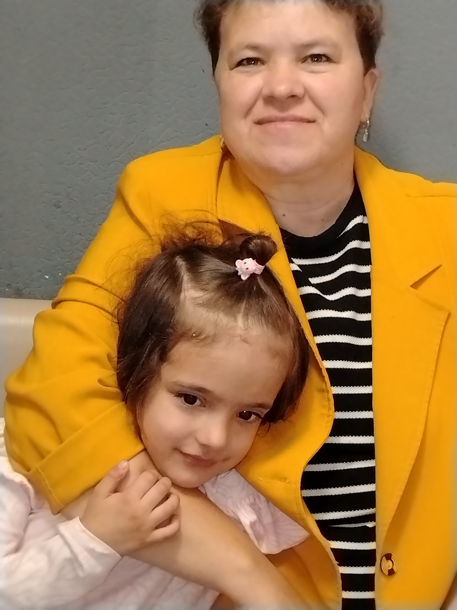 This is a medium shot photograph featuring two individuals, an adult woman and a young girl, in what appears to be an indoor setting. The woman is positioned behind the girl, embracing her. Both are facing the camera. The woman has short, dark hair and is wearing a bright yellow jacket over a black and white striped shirt. She has a gentle smile on her face. The young girl, with dark hair styled in a bun with a pink clip, is looking towards the camera with a sweet expression. She is wearing a light pink shirt. The background is a textured gray wall, suggesting an indoor environment. The lighting appears to be artificial, typical of an indoor setting. The overall mood is warm and affectionate, highlighting the bond between the two subjects. There are no visible text elements in the image. The image's context is Noviy, Russia.