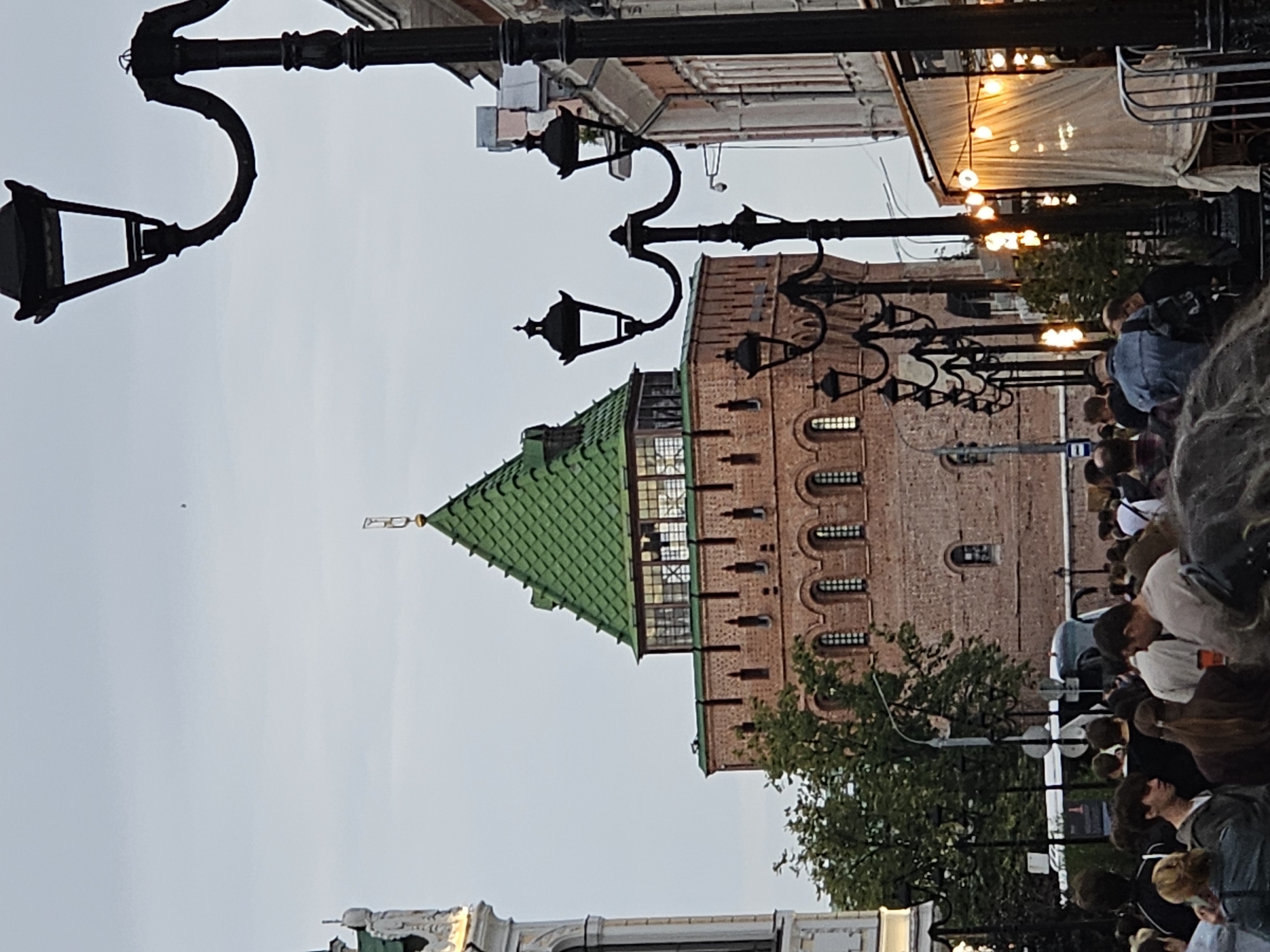 The image depicts a street scene in Nizhny Novgorod, Russia, with a prominent red brick tower with a green roof in the background. The tower features numerous arched windows and battlements. In the foreground, a crowd of people walks along a street lined with lampposts and buildings. One building on the right has outdoor seating with string lights. The sky is overcast.