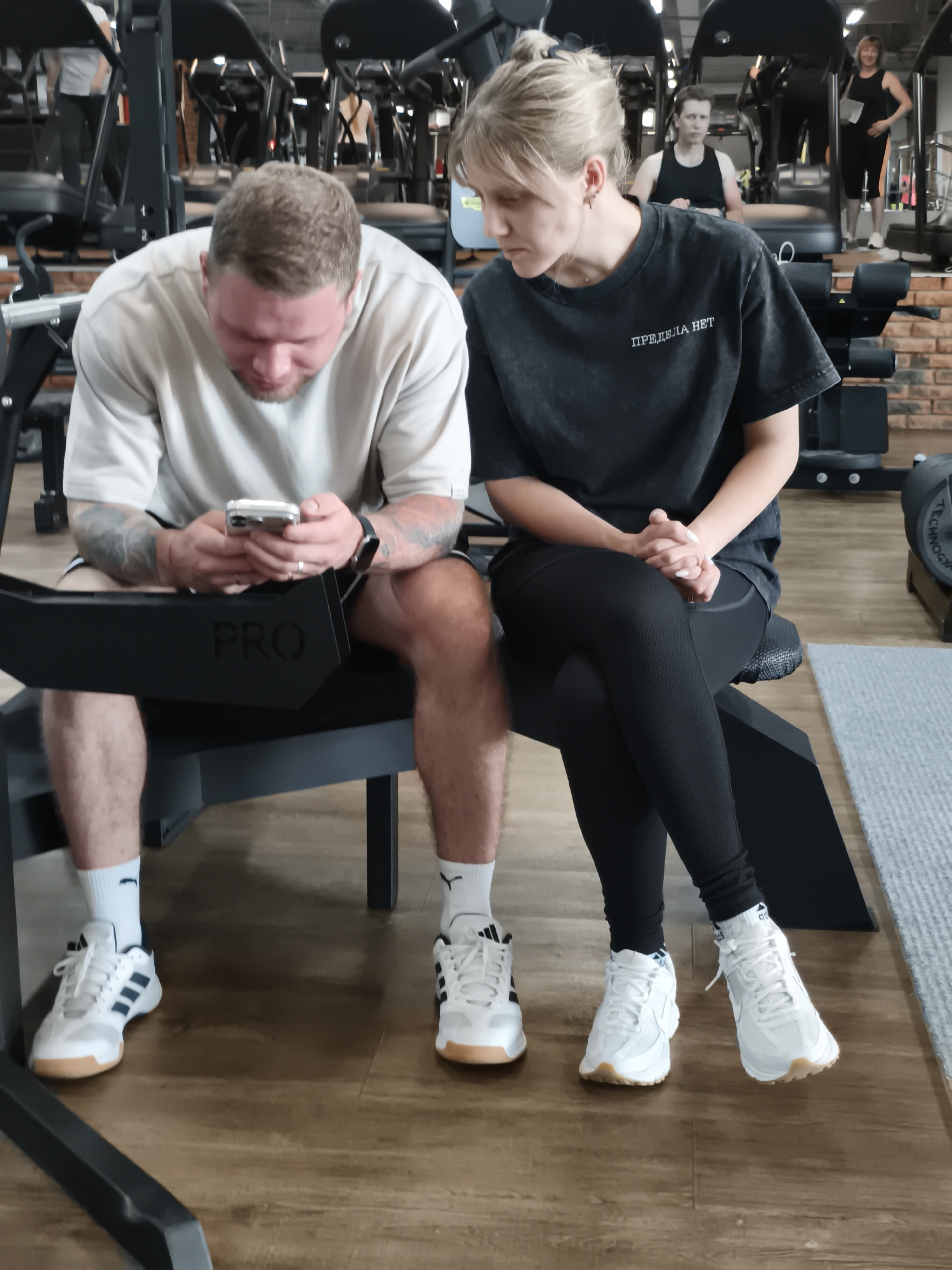 A man and a woman are sitting side by side on a piece of gym equipment indoors. The man, on the left, has short, light hair and is wearing a light-colored t-shirt, dark shorts, and white sneakers with blue stripes. He is looking down at a smartphone he holds in both hands. His arms are tattooed. The woman, on the right, has blonde hair pulled back in a bun and is wearing a dark t-shirt, black leggings, and white sneakers. Her hands are clasped in her lap, and she is looking down towards the man. The equipment they are sitting on has the word "PRO" on it in large letters. In the background, there are treadmills and other gym equipment, along with other people working out. The floor is made of wood, and there is a brick wall visible in the background. The location is Chaikovsky, Russia.