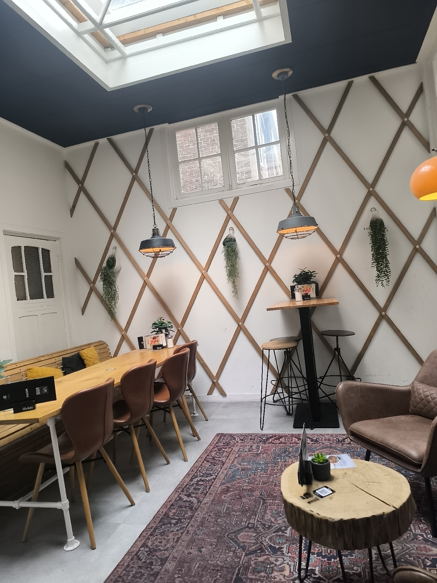 The image shows the interior of a cafe in Hoorn, Netherlands. The cafe has a modern and rustic design with a diagonal wooden lattice on the walls. The ceiling is dark with a skylight. There are several hanging plants, pendant lights, and a large wooden table with brown chairs. In the foreground, there is a round wooden table with a small plant and a menu. A brown leather armchair is visible on the right side of the image. The floor is covered with a patterned rug. There are no people visible in the image.