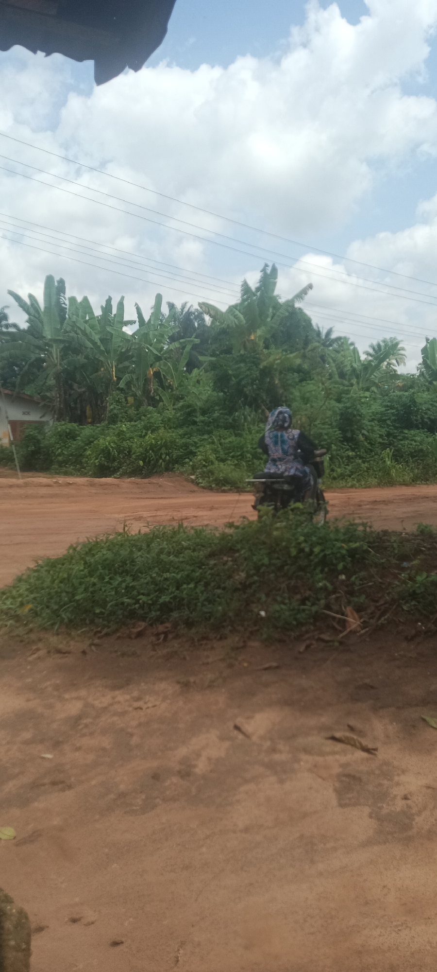 The image depicts an outdoor scene in Amauzu, Nigeria. The foreground shows a dirt road with some grassy areas. In the background, there is dense vegetation, including banana trees. A person is riding a motorcycle on the dirt road, heading away from the viewer. The sky is partly cloudy with visible power lines. A partial view of a dark object, possibly a building overhang, is in the upper left corner of the frame.