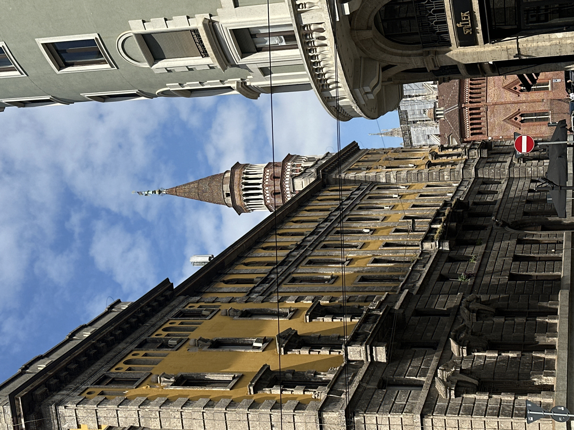 The image shows a street scene in Milan, Italy, with a view looking upward towards several buildings. The Torre Velasca, with its distinctive mushroom-like shape and a spire topped by a statue, is prominently featured. The tower is visible between other buildings, including a light grey building with windows above and a building with a sign that reads “STILLER." The buildings show signs of traditional architecture, with detailed facades, many windows, and decorative elements. A “No Entry” sign is seen. The sky is partly cloudy and blue. There are power lines intersecting the view. The photograph is taken from a low angle, emphasizing the verticality of the buildings.