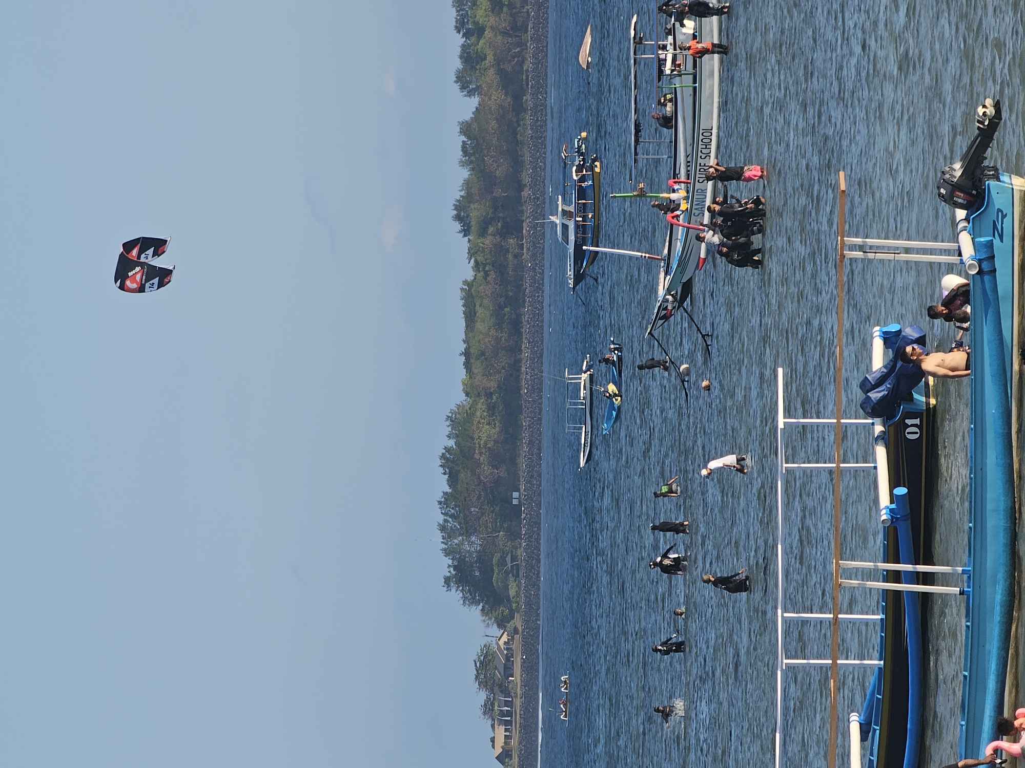 The image depicts a sunny day at a beach in Denpasar, Indonesia, with several people engaged in various water activities. The scene is divided into distinct layers, from the sky to the sea and the shoreline.
In the sky, a colorful kite with red, white, and black sections, bearing the branding "Rebel" and the number "14", is prominently visible.
The sea is populated with numerous individuals, many of whom appear to be wading or swimming in the shallow waters. Some are dressed in dark wet suits, suggesting diving or surfing activities. A group of people is gathered near a boat labeled "SANUR SURF SCHOOL". Several boats are present in the water, including a larger one with a canopy, possibly a dive boat, and smaller outrigger boats. One boat in the foreground has the number "01" painted on its side.
In the background, a tree-lined coastline forms the horizon, with a few buildings nestled among the trees. The water is calm, reflecting the clear blue sky. The overall atmosphere suggests a recreational and active environment, likely a popular tourist spot.