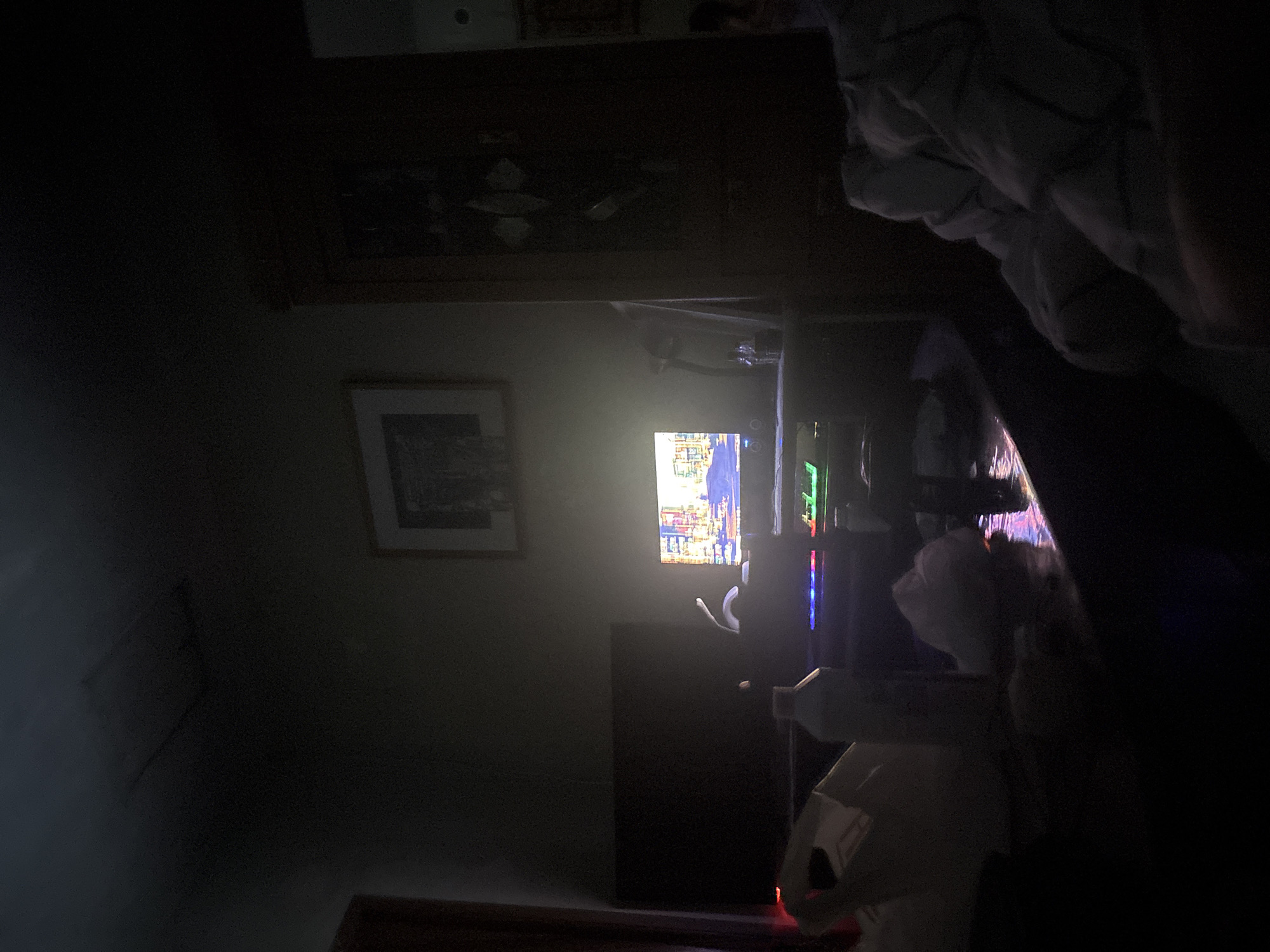 The image depicts a darkened room with a monitor displaying a colorful scene, possibly a video game, as the primary light source. To the left of the monitor, a framed picture is visible on the wall. Beneath the monitor, a dark object, potentially a speaker or another electronic device, is situated on the floor. In the lower right corner, there's a partial view of a white object. The scene appears to be indoors and illuminated primarily by the monitor's light. It is not possible to determine if this image depicts Yerevan, Armenia, or to infer any specific details about people, subjects, actions, or interactions based on the information in the image.