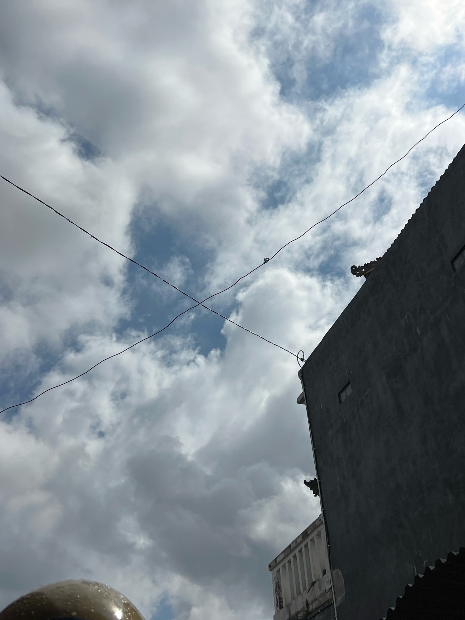 The image shows a cloudy sky with patches of blue. Two thin wires are visible, crossing each other in the frame. In the foreground, the upper portion of a dark gray building is visible, with decorative architectural elements at the top. The building has a textured, concrete-like surface and a dark, corrugated roofline. There are also some small rectangular windows on the building's facade. The overall impression is an urban scene in Denpasar, Indonesia, with a focus on the sky and the architecture. The lighting suggests it is daytime, possibly late afternoon or early morning, with the sun partially obscured by clouds.