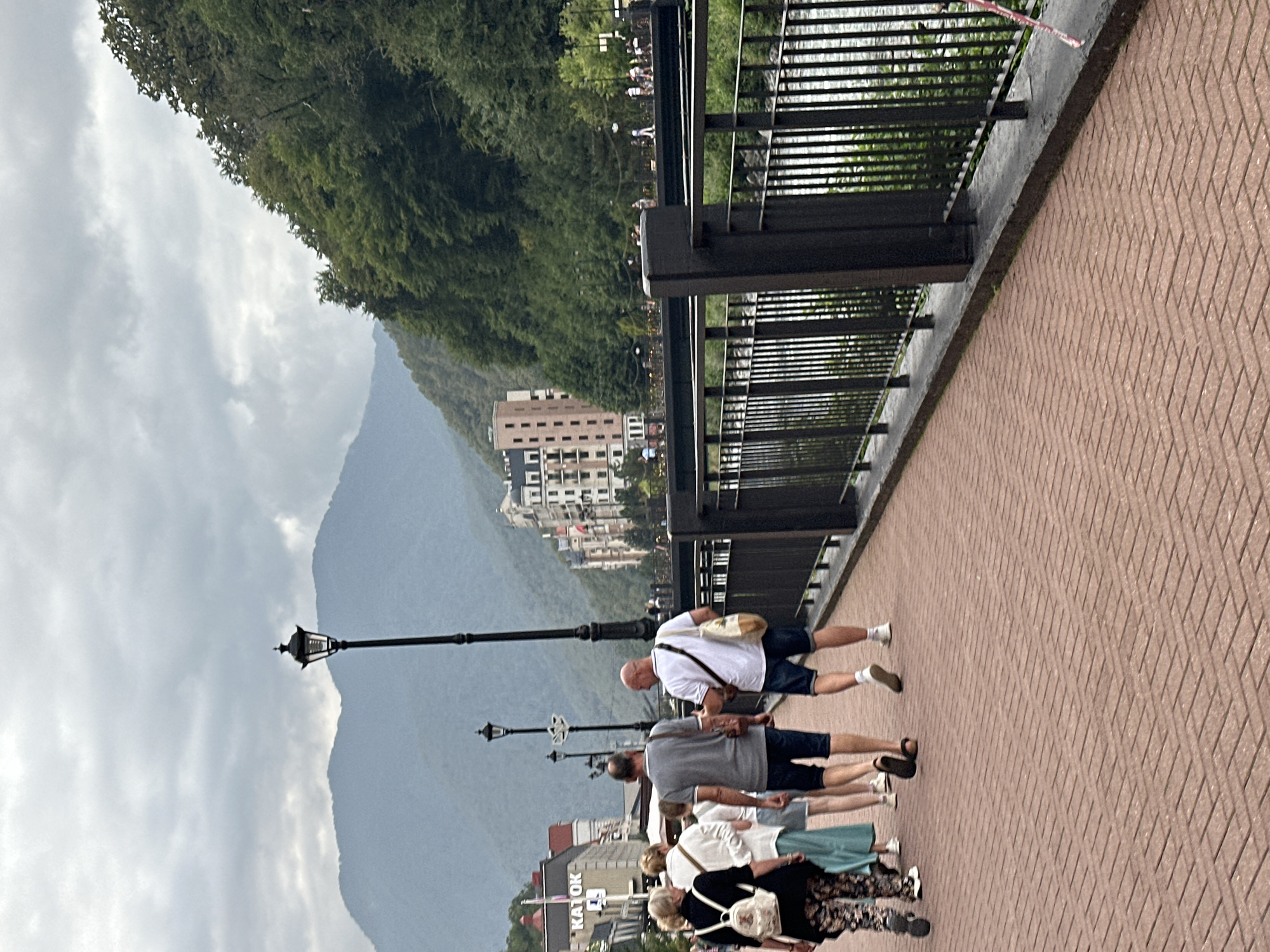 This is a full-shot image taken outdoors on a cloudy day in Estosadok, Russia. The foreground shows a brick-paved walkway with a group of people walking away from the camera. On the left, a woman with blonde hair wears a black top with a patterned skirt and a white backpack. Next to her, a woman with blonde hair wears a white cardigan over a light blue skirt. Behind them, a woman in a light-colored top and blue skirt walks. Two men are in the center foreground, both walking away. One man wears a gray t-shirt and dark shorts, with a brown bag slung over his shoulder. The other man is bald and wears a white t-shirt and dark shorts, with a beige bag over his shoulder.
To the right of the walkway is a black metal railing overlooking a body of water with a visible current. Beyond the railing, there is a bridge with people walking on it. In the background, there are buildings with European-style architecture, ranging from multi-story hotels to smaller establishments, some with red roofs. Behind the buildings, a dense forest covers a steep mountainside. The furthest background consists of hazy, layered mountains under a cloudy sky. A lamppost is visible on the walkway in the mid-ground, and another is partially visible in the far background. The overall scene suggests a resort or tourist area.