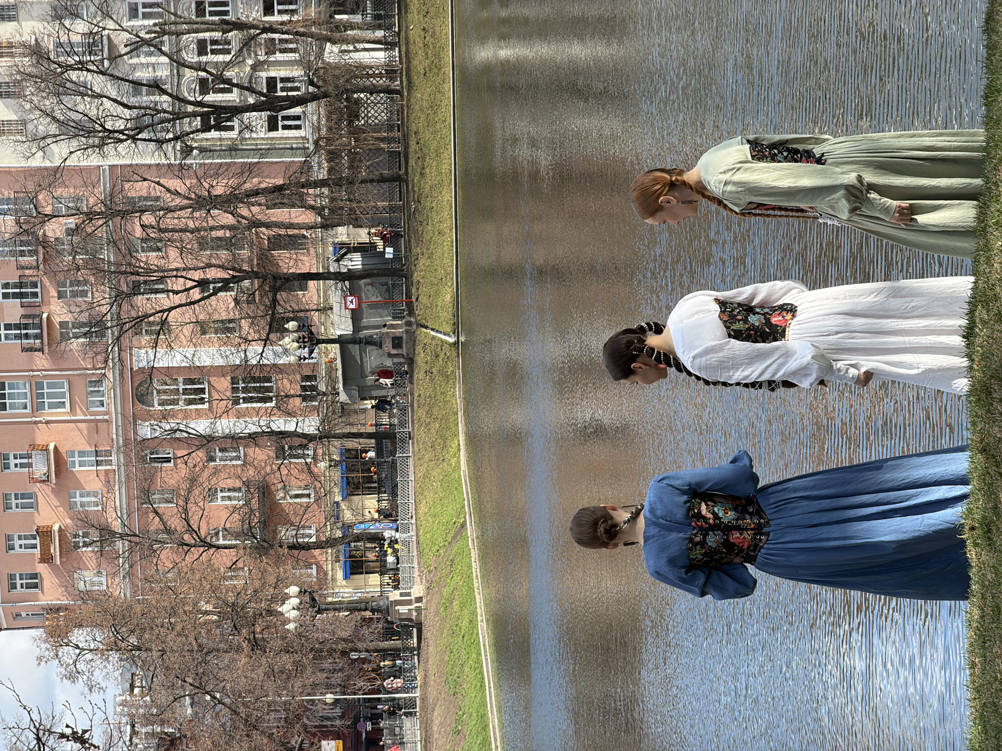 Three women stand near a body of water. The woman in the foreground is wearing a long, light blue dress with a dark floral corset. The next woman is wearing a similar style of dress but in white, and the third woman is in a light green dress, also with a dark floral corset. All the women have their heads bowed, looking towards the water. The water appears to be a pond or canal with reflections of the sky and surrounding buildings.
In the background, across the water, is a street with people, trees, buildings, and cars. The buildings are of a classic design, with multiple windows and a peach-colored exterior.
The image is taken in Moscow, Russia, in the area near the Patriarch's Ponds.