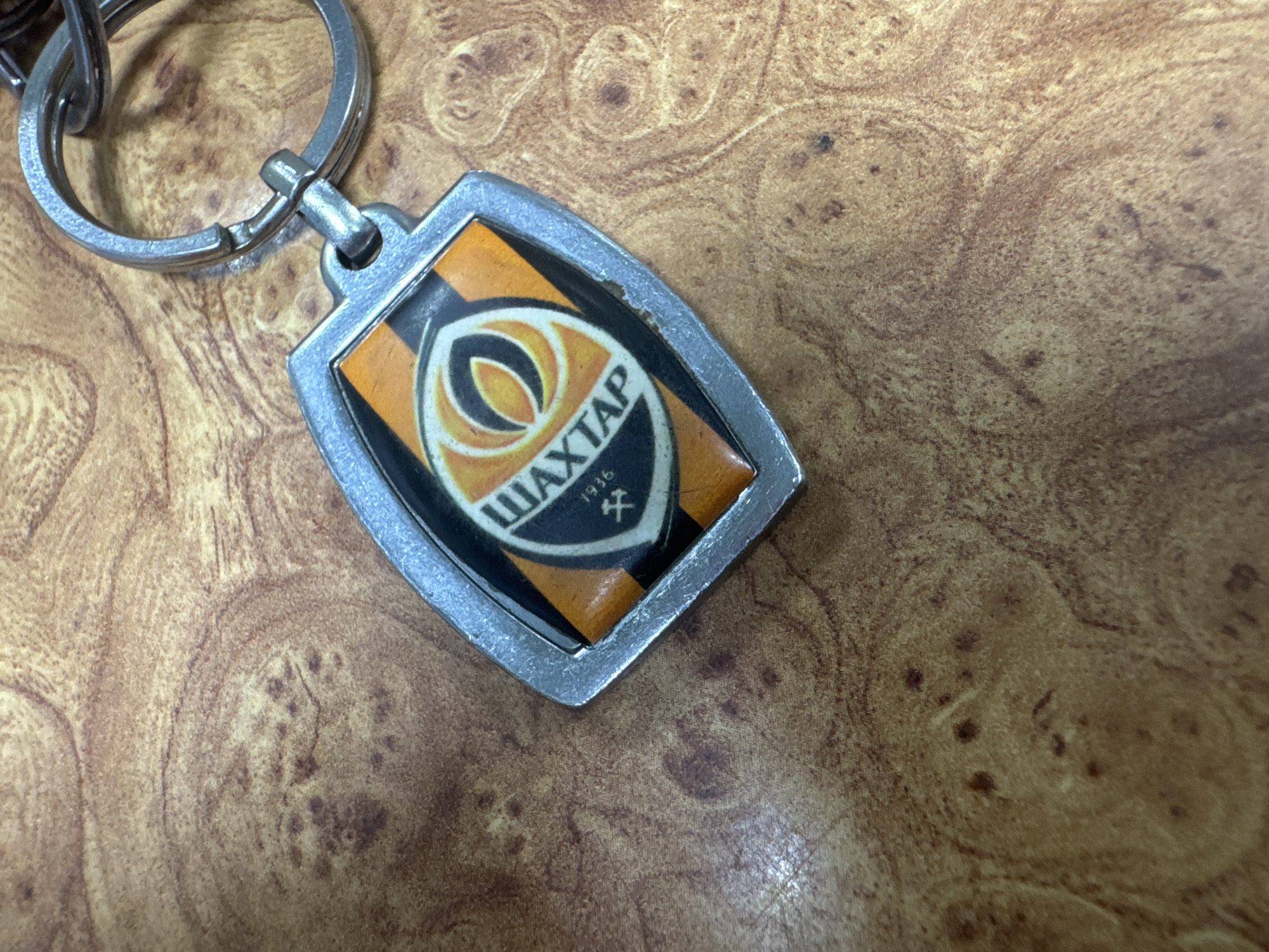 A close-up shot shows a silver keychain with a shield-shaped emblem attached to a metal ring. The emblem features the logo of FC Shakhtar Donetsk, a Ukrainian professional football club. The logo is divided into orange and black stripes, with a white oval containing a stylized yellow flame or leaf-like symbol. The word "ШАХТАР" is written in Cyrillic script above the symbol, and "1936" is printed below it, along with a small, black, crossed-pickaxe symbol, referencing the club's mining origins in Makiivka, Ukraine. The keychain rests on a textured, wood-like surface with a swirling grain pattern.