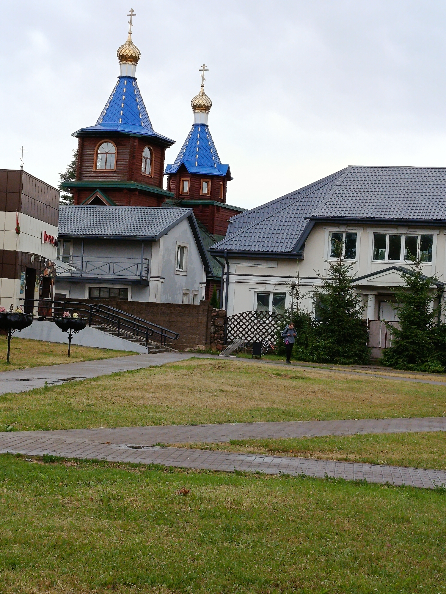 The image shows a complex of buildings under a cloudy sky. In the background, there are two distinct church structures with golden domes and bright blue roofs, topped with crosses. To the left, a portion of a modern brown and white building is visible. In the middle ground, a light-colored, two-story house with a dark grey tiled roof stands prominently. To the right of this house, several evergreen trees are planted. A person, wearing a grey jacket and dark pants and carrying a pink bag, stands on a paved path in front of the house, near a black decorative fence and a bicycle. The foreground consists of a grassy lawn with paved walkways. The location is identified as Смілавіцкі сельскі Савет, Belarus.