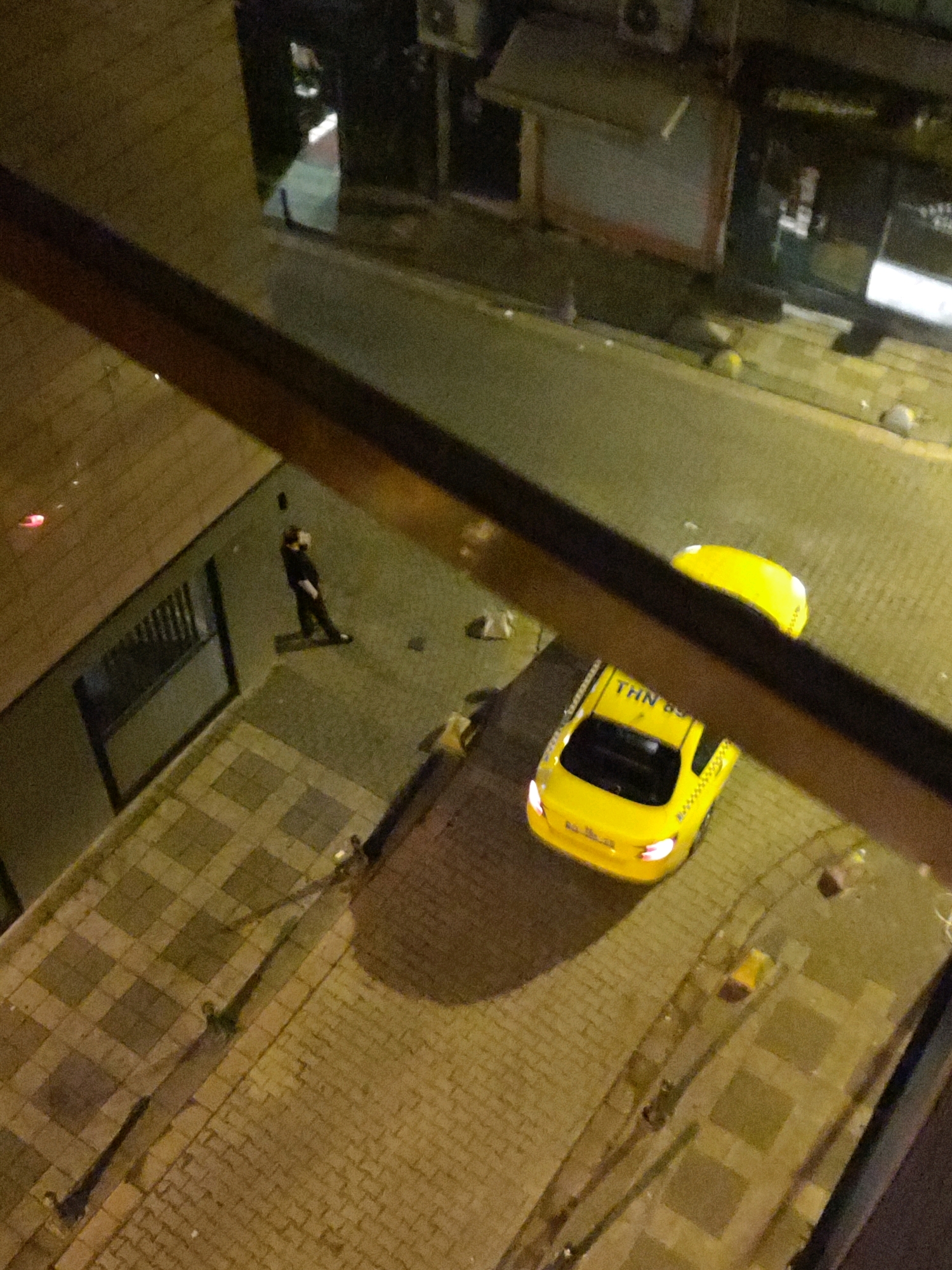An aerial view captures a street scene at night in Kadıköy, Turkey. A person dressed in dark clothing stands on a paved walkway next to a building. Across the street, a yellow taxi is parked, its headlights illuminated. The street and walkway are made of interlocking pavers. The image is taken from an elevated position, looking down at the scene, with a wooden railing in the foreground partially obscuring the view.
