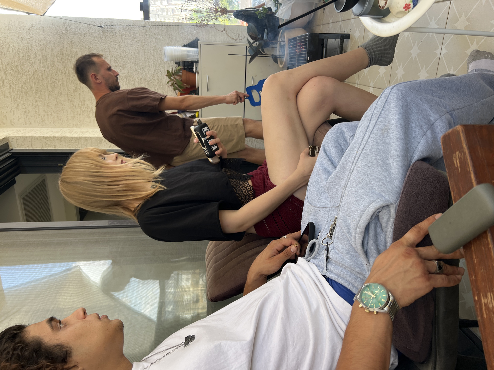 The image depicts an outdoor scene, likely a balcony, in Kiryat Bialik, Israel, given the context. Three individuals are present. In the foreground, a man wearing a white t-shirt, grey sweatpants, and a green-faced wristwatch is seated and looking to his left. His hand rests on his lap, holding a phone, with keys and a lighter nearby. To his right and slightly behind him, a woman with blonde hair, wearing a black top and a burgundy crocheted skirt, is seated with her legs crossed. She is holding a black and silver object in her right hand. Further back, a man in a brown t-shirt and khaki shorts is standing and looking downwards. He is holding a blue object. Behind him, a white cabinet is visible, along with potted plants and a small grill with charcoal. The environment suggests a casual gathering on a warm day.