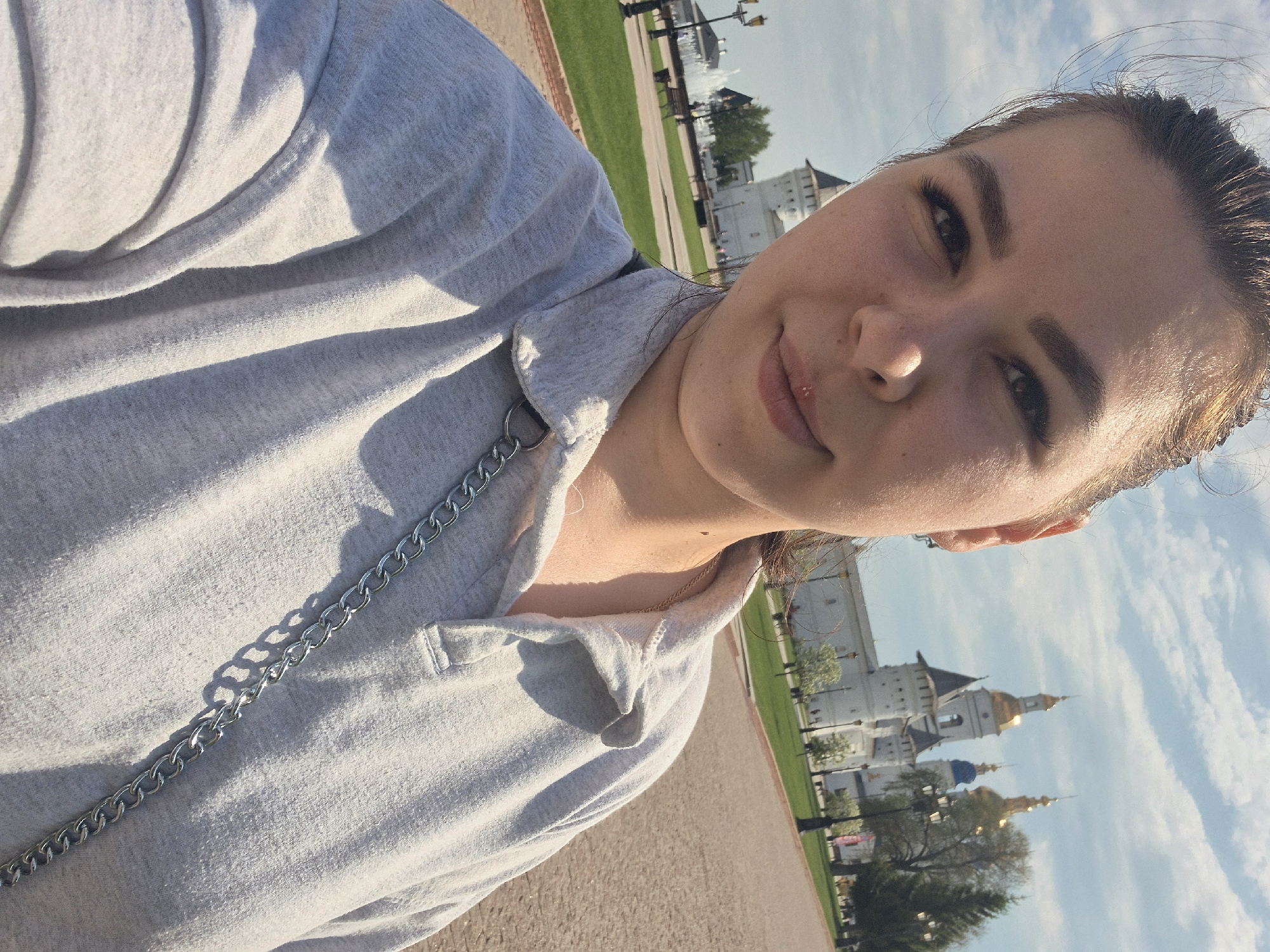The image shows a selfie of a woman. She is fair-skinned with dark hair pulled back and is wearing a light gray collared shirt with a silver chain. In the background, a building with a golden and blue dome is visible, specifically the Tobolsk Kremlin, located in Tobolsk, Russia. There are lampposts and patches of green grass in the background, and the sky is light blue with scattered clouds. The photo is taken outdoors during daylight.
