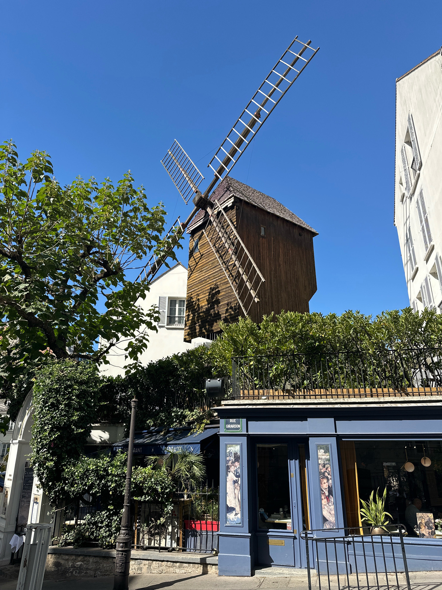 The image captures a sunny day in Montmartre, Paris, France, with a prominent wooden windmill dominating the skyline. The windmill's weathered texture and large, slatted blades are clearly visible against a bright blue sky. In the foreground, a blue building with large windows displays framed artwork, and a sign reads "RUE GIRARDON." Lush green foliage surrounds the building, and a black metal railing runs along its rooftop. A vintage lamppost stands on the sidewalk. The scene suggests a vibrant street with a mix of historical architecture and modern establishments.